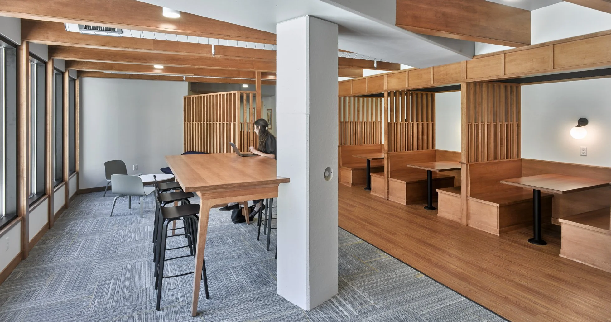 Brightly lit apartment community common space at Ella Apartments with soft seating and booths. A person is seated at a table using a laptop.