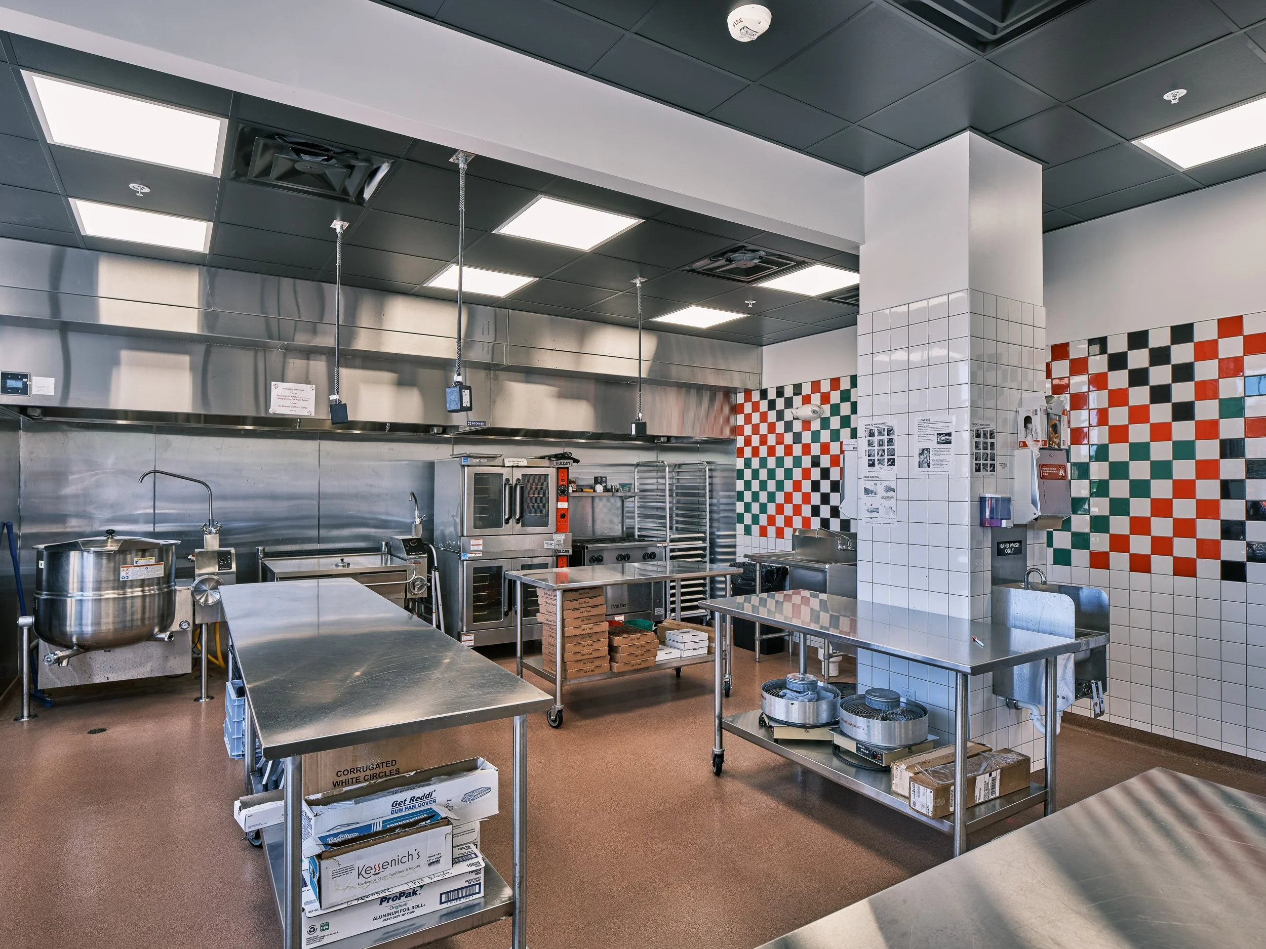 Commercial kitchen with stainless steel appliances and worktables. A black, red, green and white tile mural forms an abstract checkerboard pattern on the back wall.