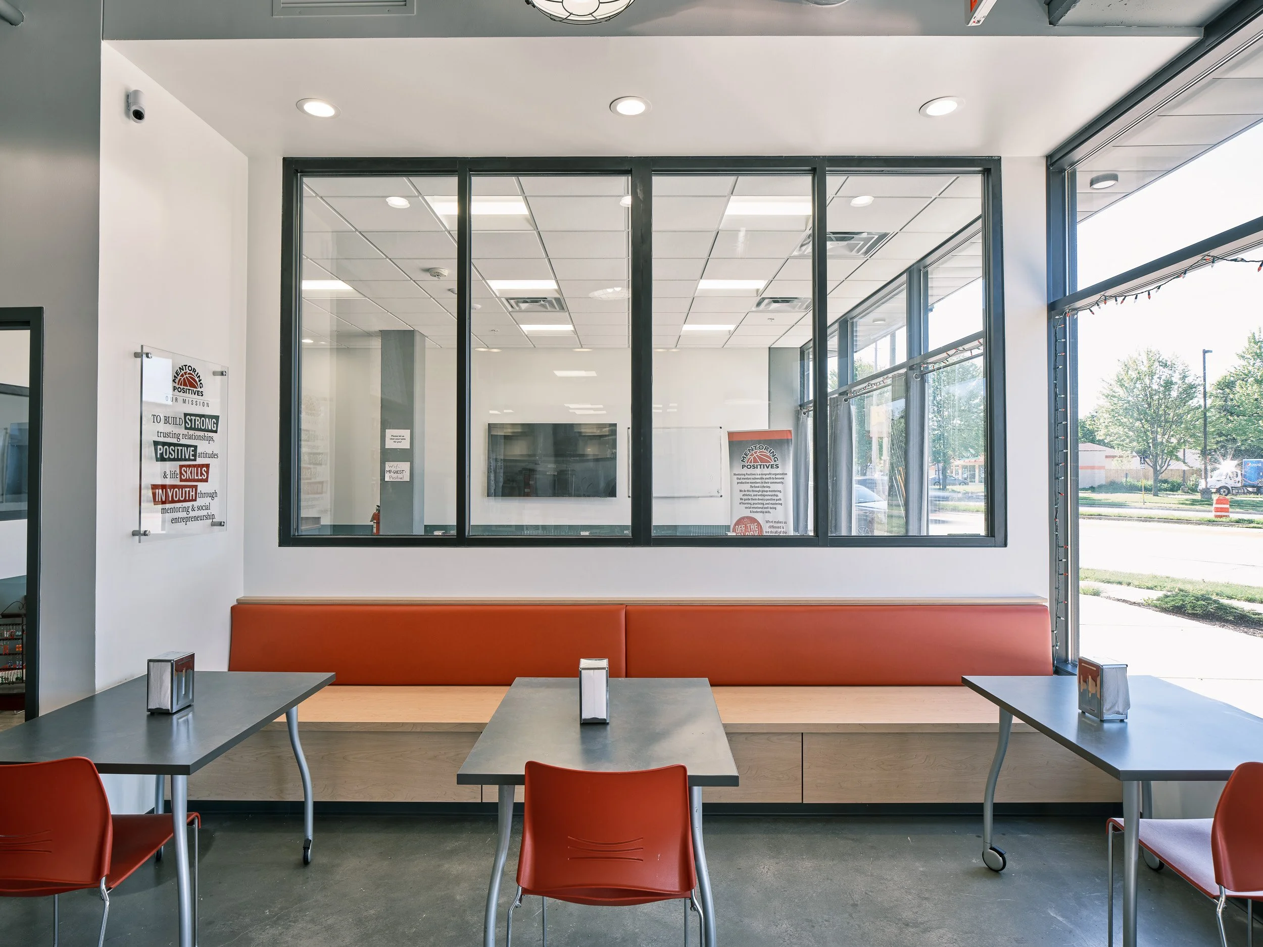 A long banquette defines the dining space with movable dining tables in the foreground. Windows look through to a multi-use space with a television and out to East Washington Avenue.