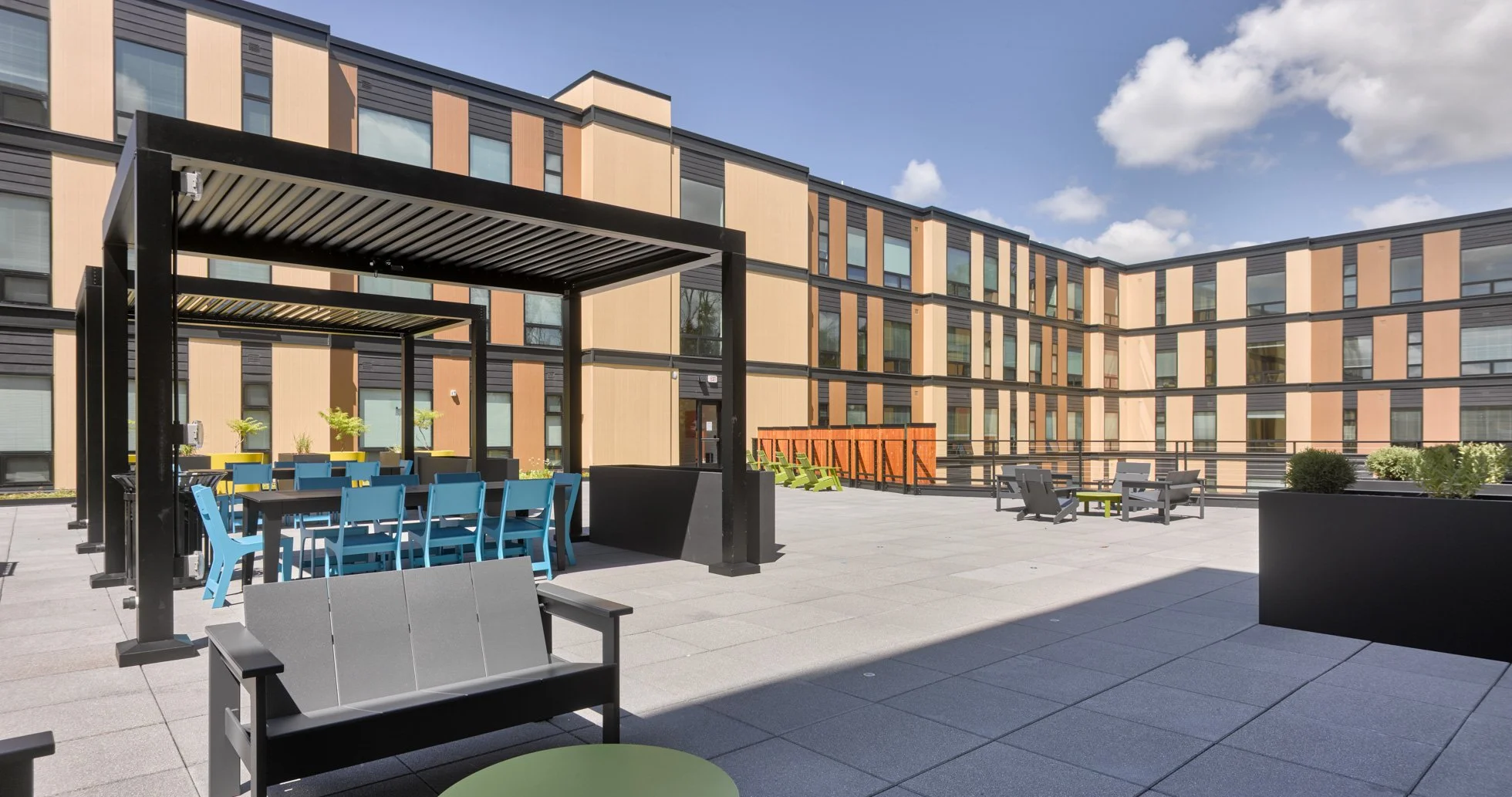 Landscaped rooftop terrace with brightly colored outdoor furniture. Pergolas shade a dining area and a modern apartment building, Ella Apartments, wraps around the terrace. 