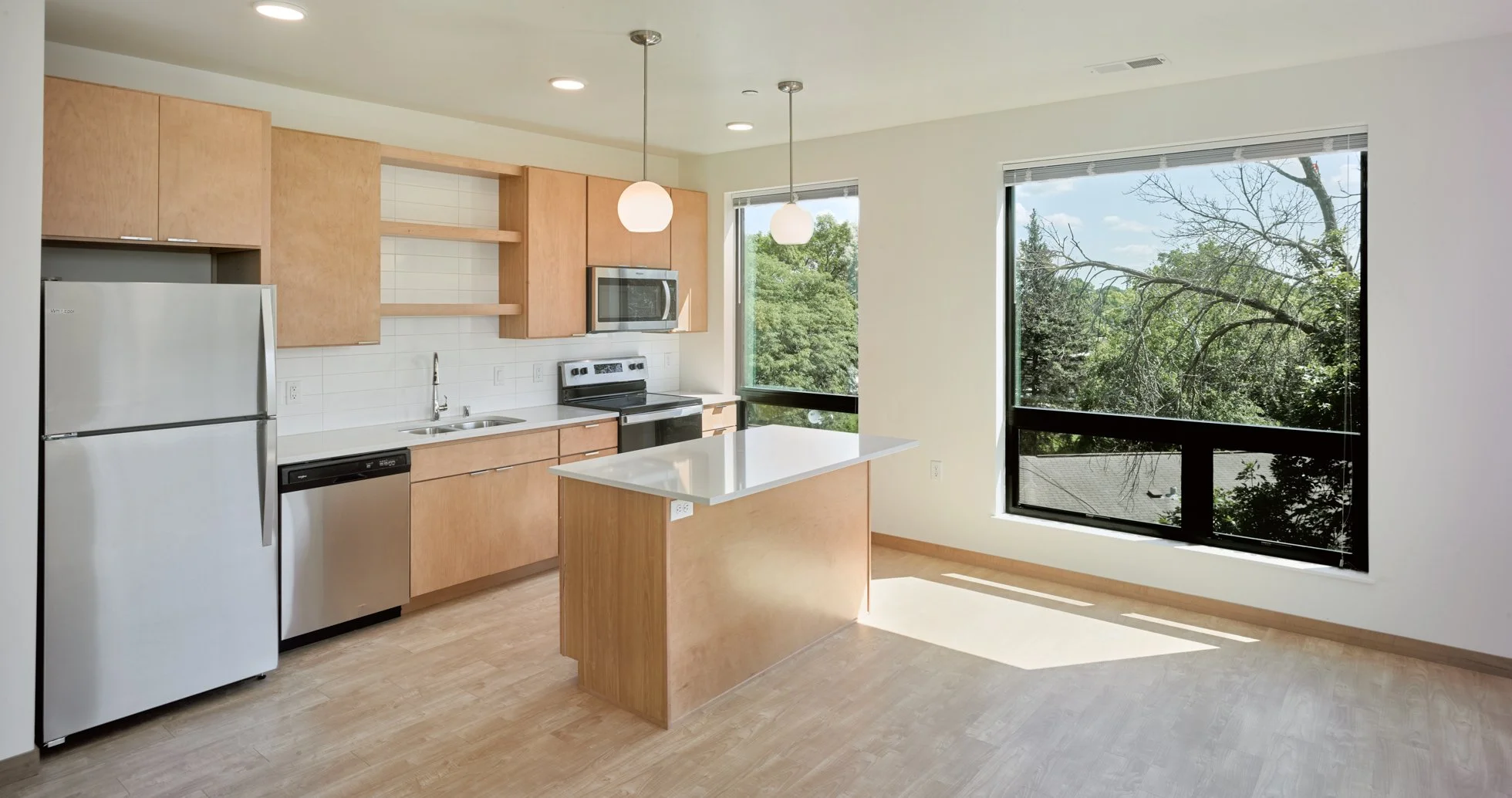 View of a kitchen inside an apartment at Ella Apartments. Light maple cabinetry and flooring paired with stainless steel appliances and white countertops provide airy surroundings for residents to call home. 