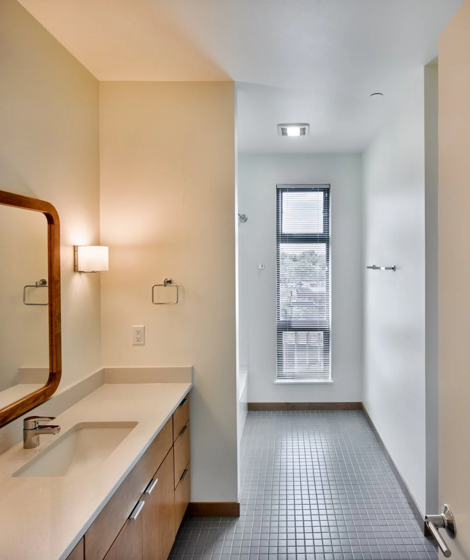 View of a bathroom vanity in Ella Apartments. Maple cabinetry and white countertops are accented by a handmade, rounded corner, rectangular mirror made from chunky wood. Light floods in from the window.