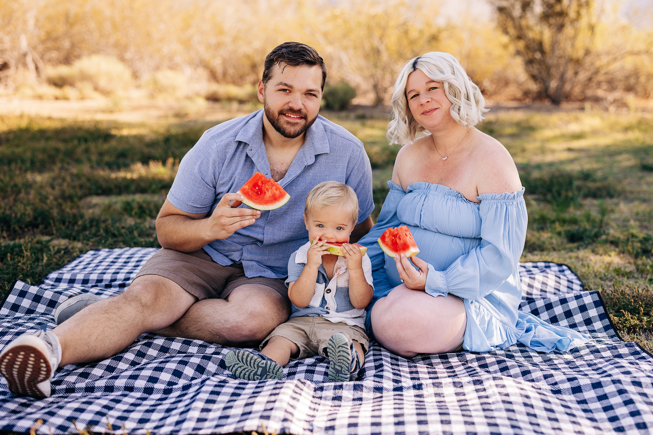 A family of three sitting on a checkered picnic blanket outdoors, eating watermelon slices, with trees in the background.
