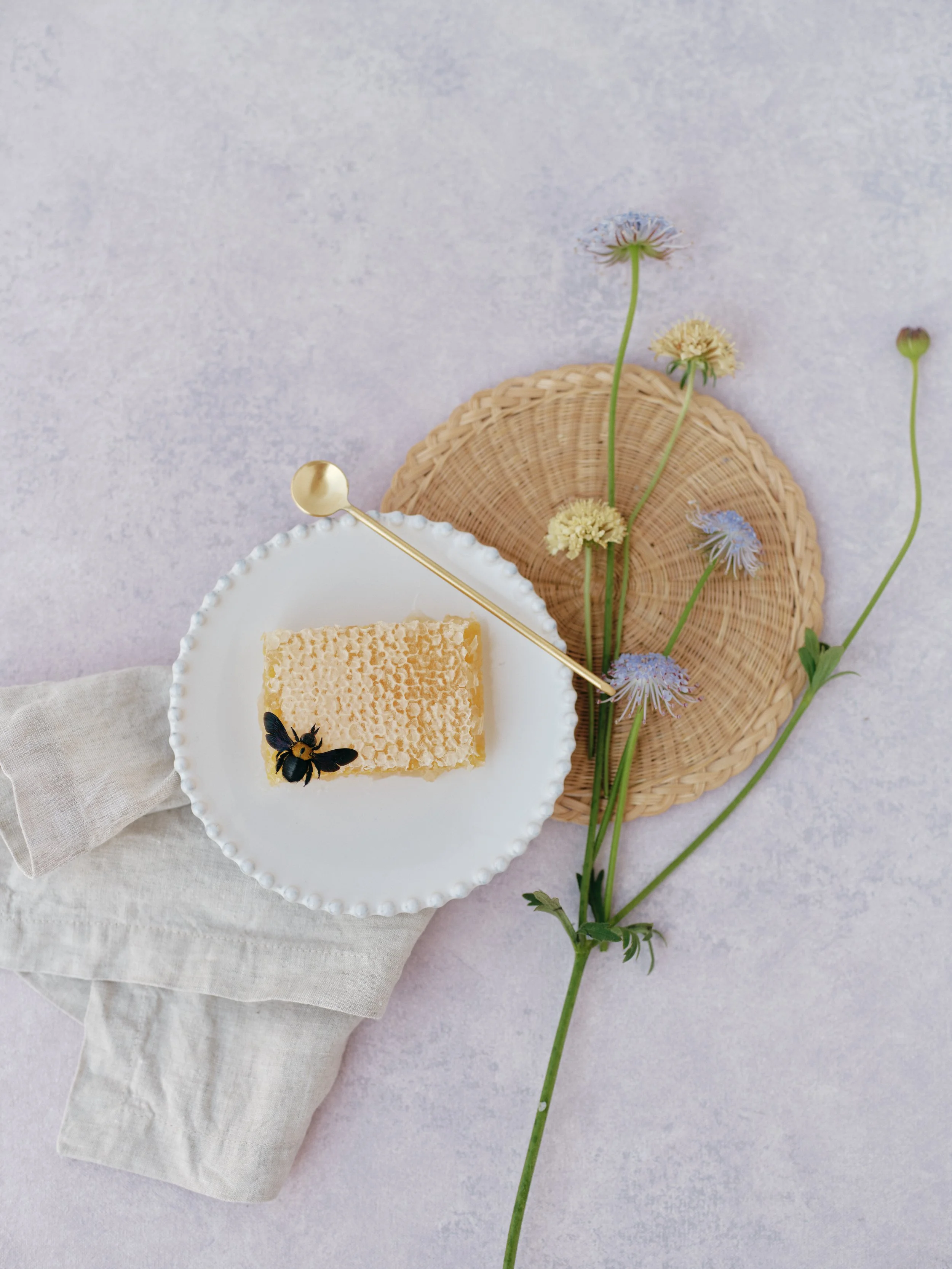 Honeycomb on a bee-illustrated plate with gold spoon, scabiosa flowers, and woven tray styled on a soft lavender hand-painted surface