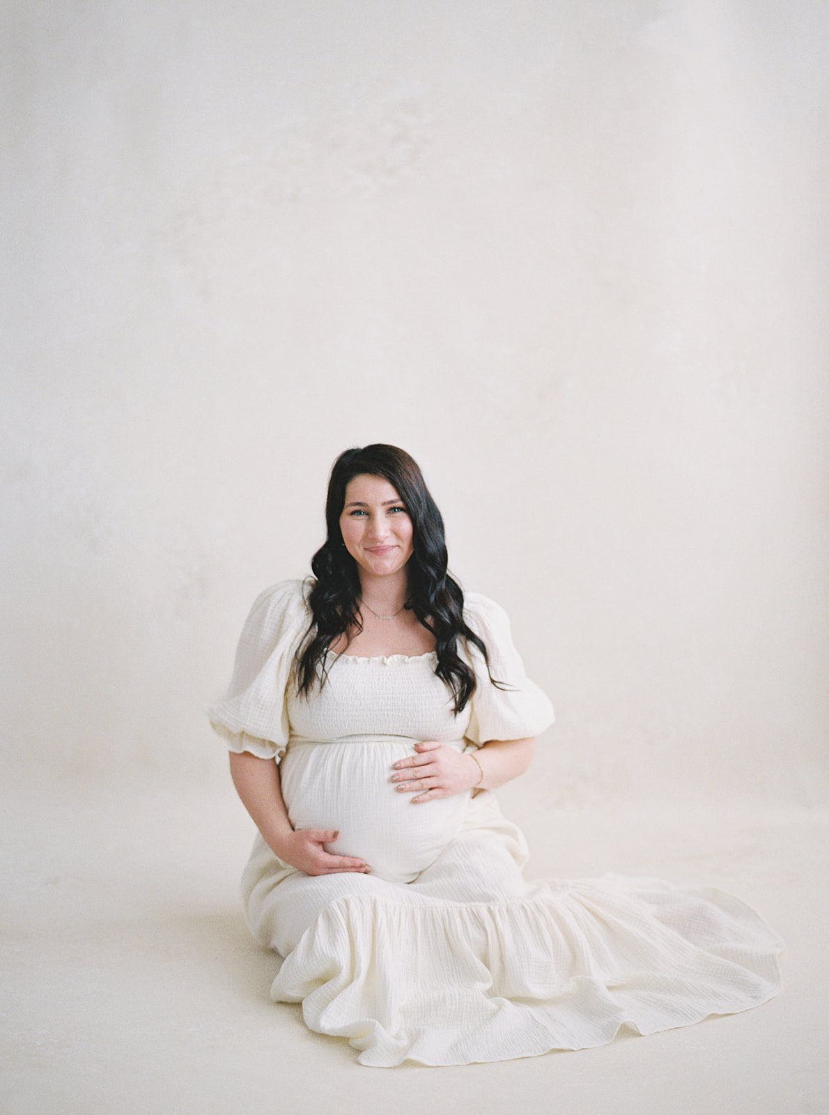 expecting mother in white gown seated on a Chasing Stone Limestone hand-painted canvas backdrop