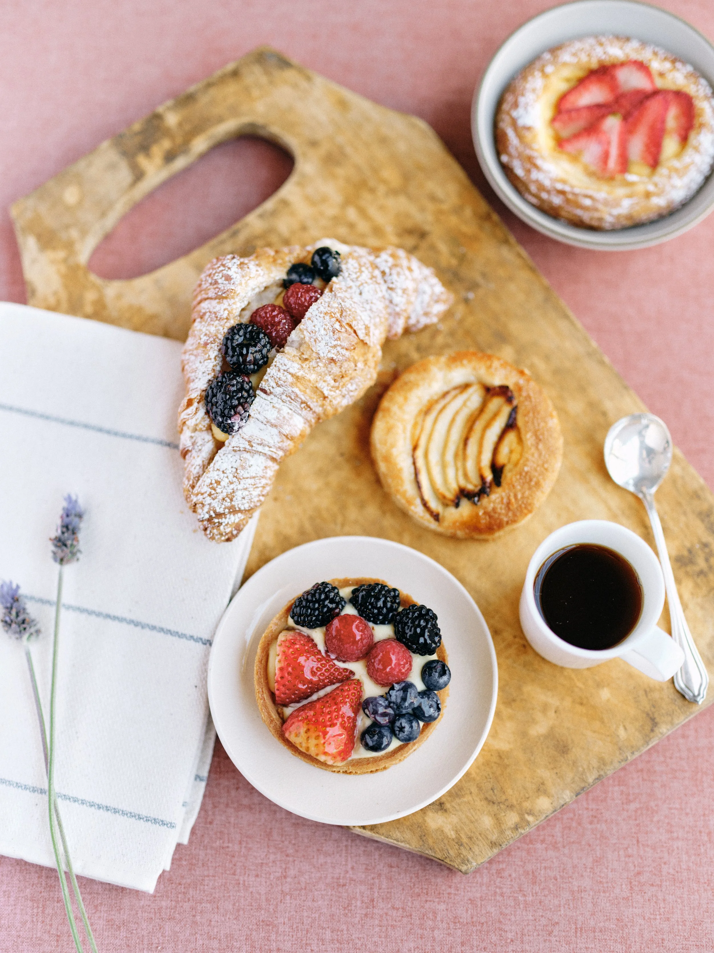breakfast spread flat lay with berry croissant, fruit tarts, and espresso on a wooden cutting board styled on a warm blush hand-painted surface