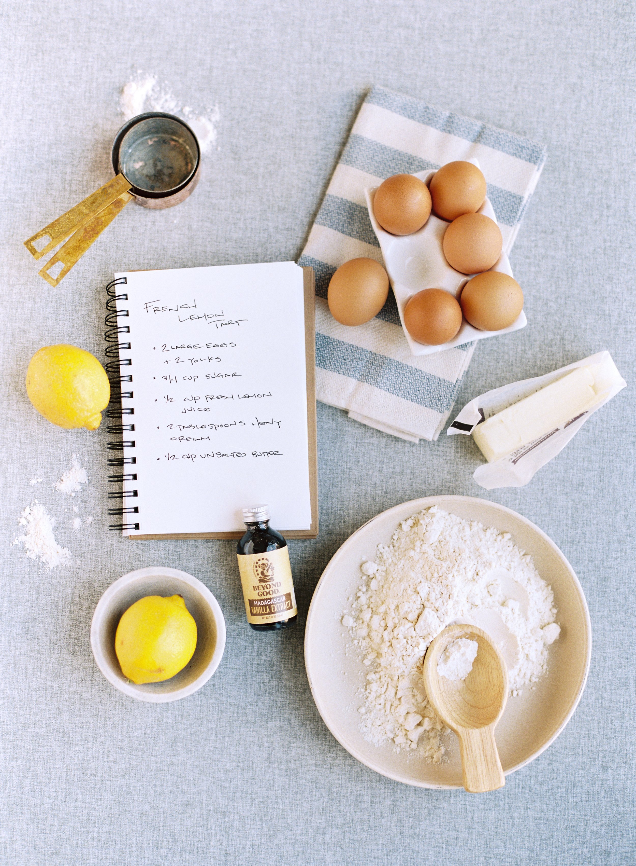 Balanced flat lay composition of French lemon tart ingredients on cool-toned fabric styling surface with recipe notebook, eggs, lemons, and baking supplies arranged with generous negative space