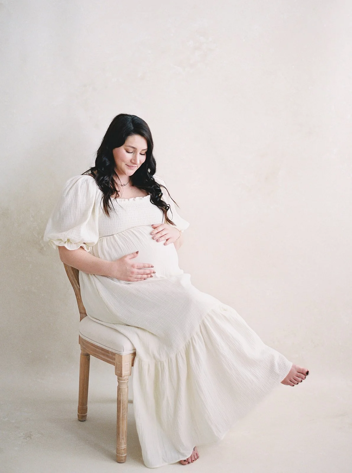 pregnant woman in cream dress seated against a Limestone hand-painted canvas backdrop, demonstrating 70/30 negative space maternity portrait composition