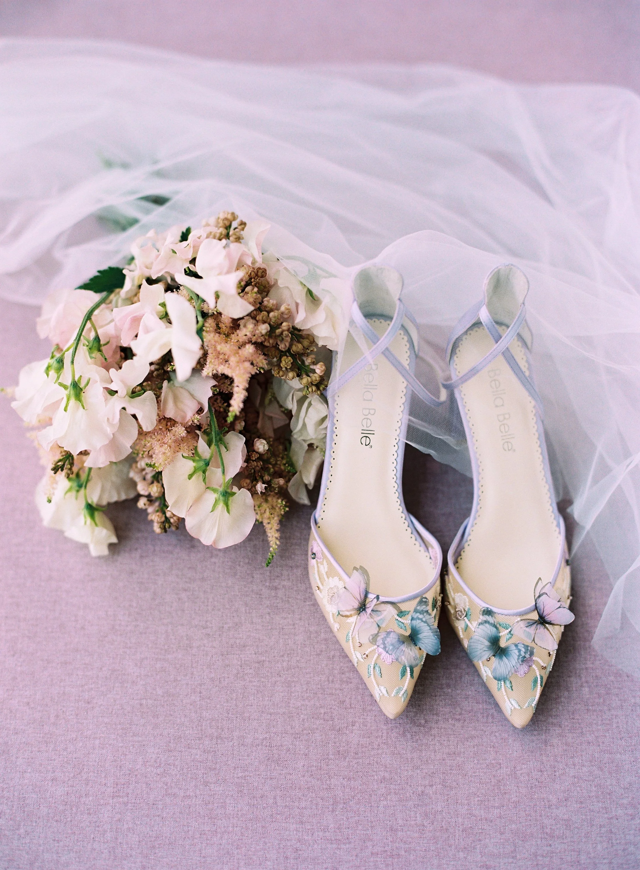 Romantic wedding flat lay with white and blush bouquet, embroidered butterfly shoes, and flowing veil styled on lavender gray surface