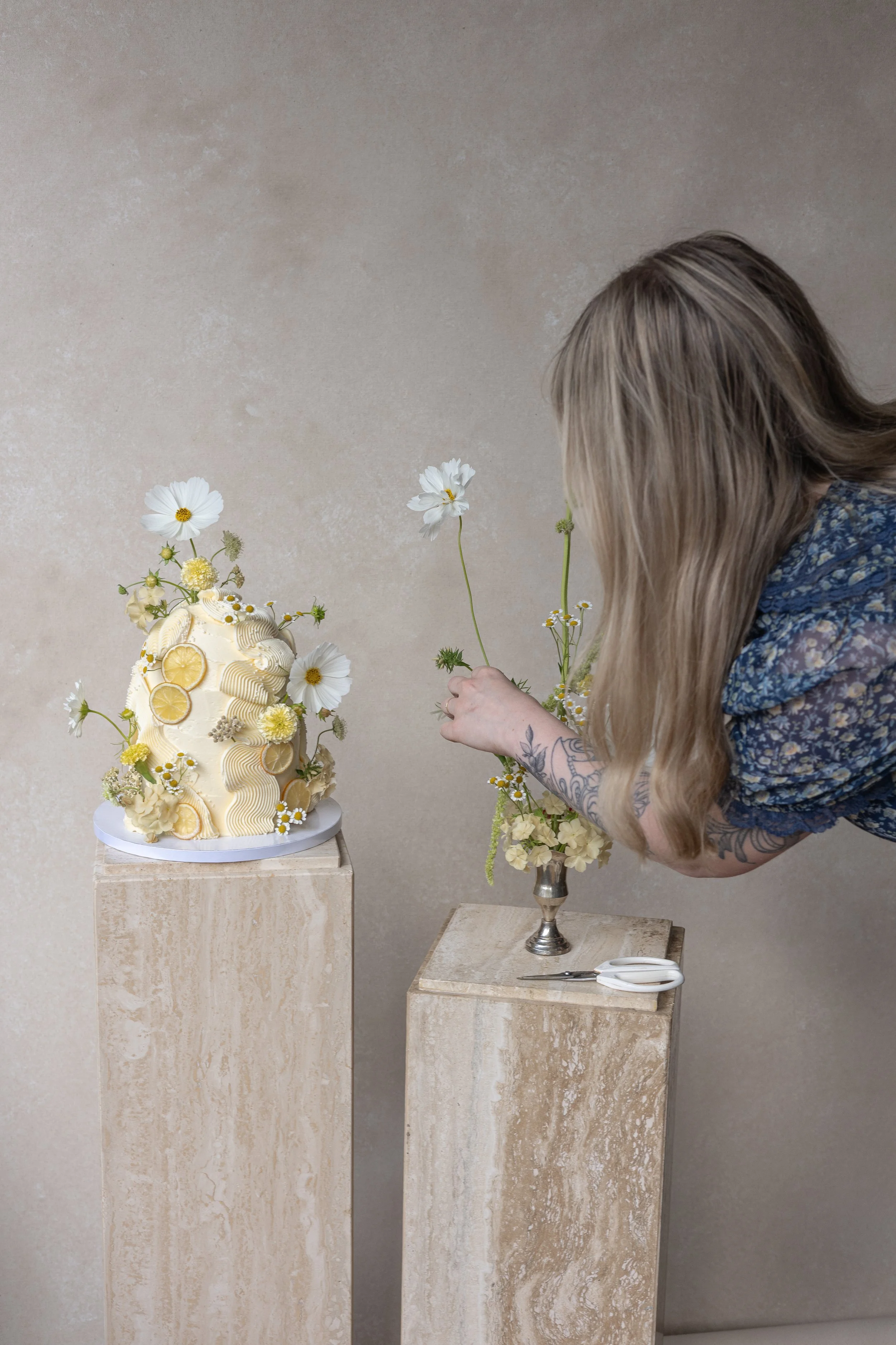 Woman styling a floral-decorated wedding cake with lemon slices on a neutral textured backdrop, arranging fresh flowers beside the cake on stone pedestals