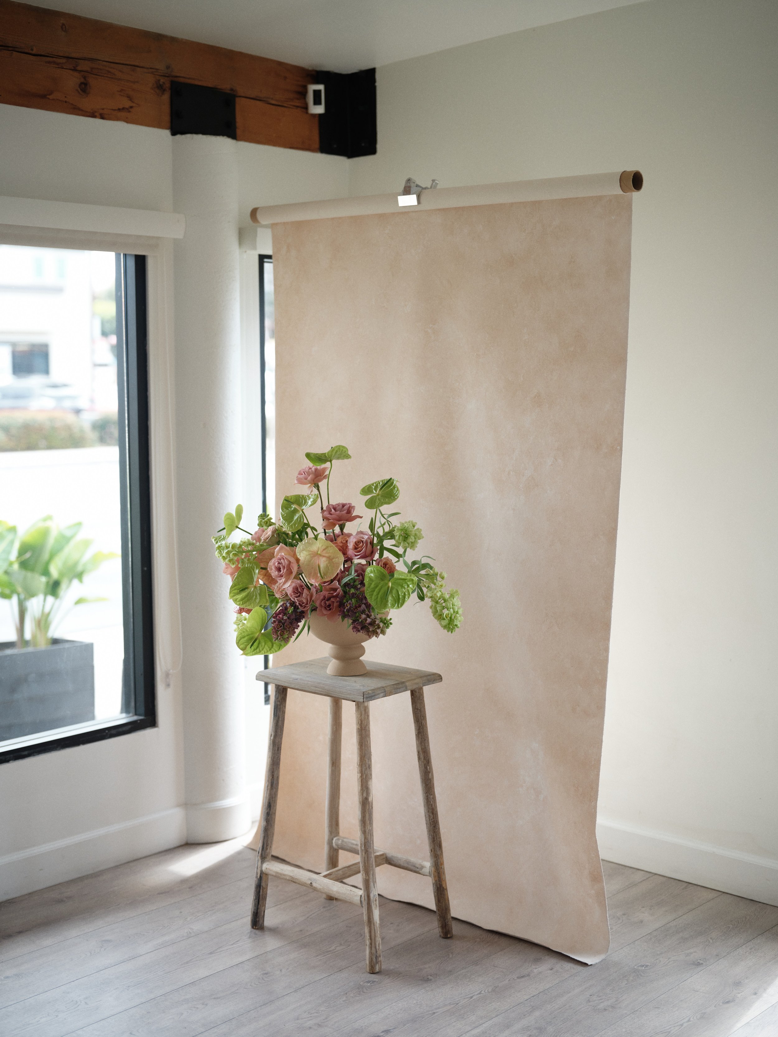 warm cream hand-painted canvas backdrop hanging in a studio with a pink rose and anthurium floral arrangement on a rustic wooden stool