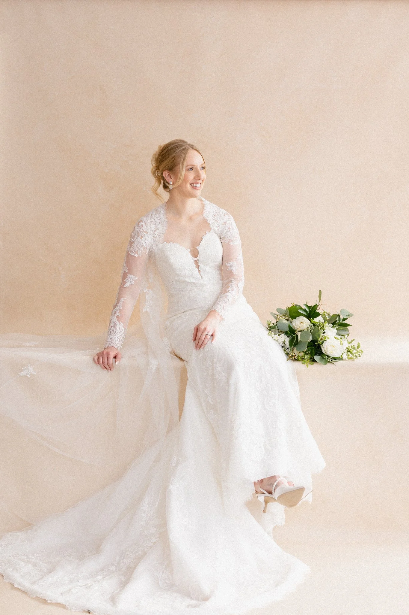 Bride in lace wedding dress sitting on neutral studio backdrop holding bouquet, full-length bridal portrait