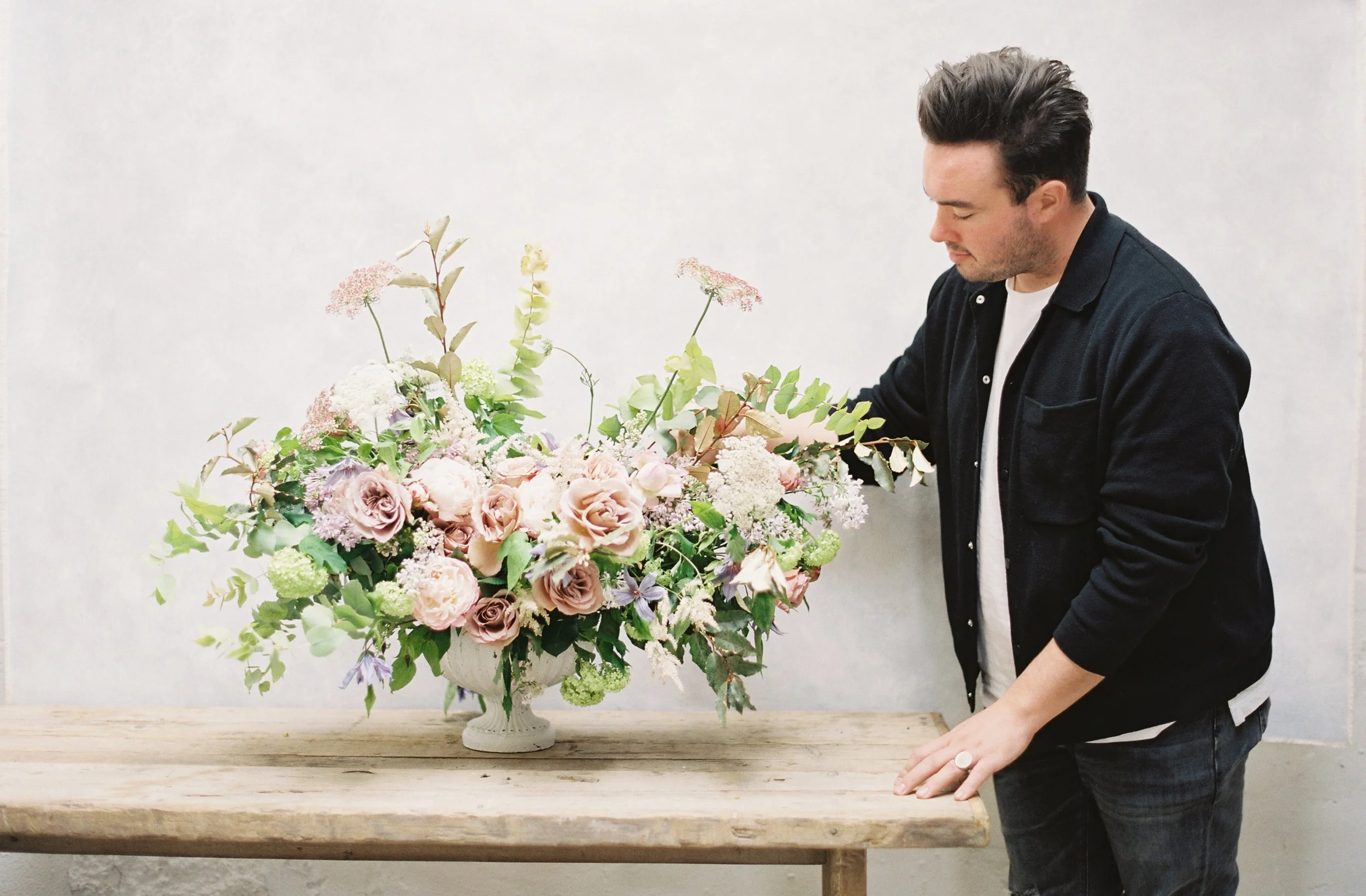 Photographer styling a large pastel floral arrangement on a rustic wooden table against a neutral textured backdrop, demonstrating creative backdrop composition techniques
