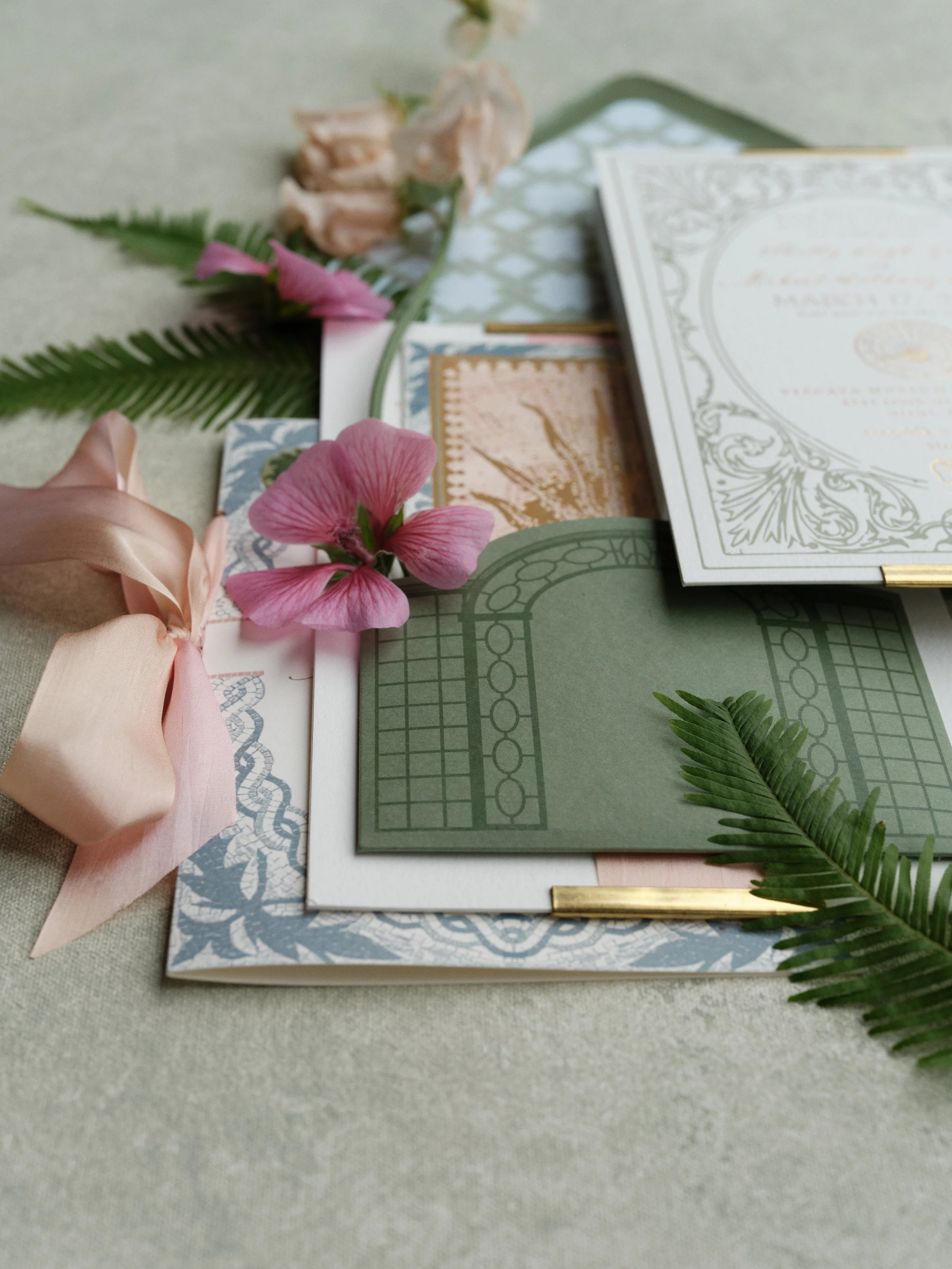 Wedding invitation suite gathered flat lay composition with layered stationery, blush ribbon, pink geranium, and tropical fern accents on textured sage green photography surface