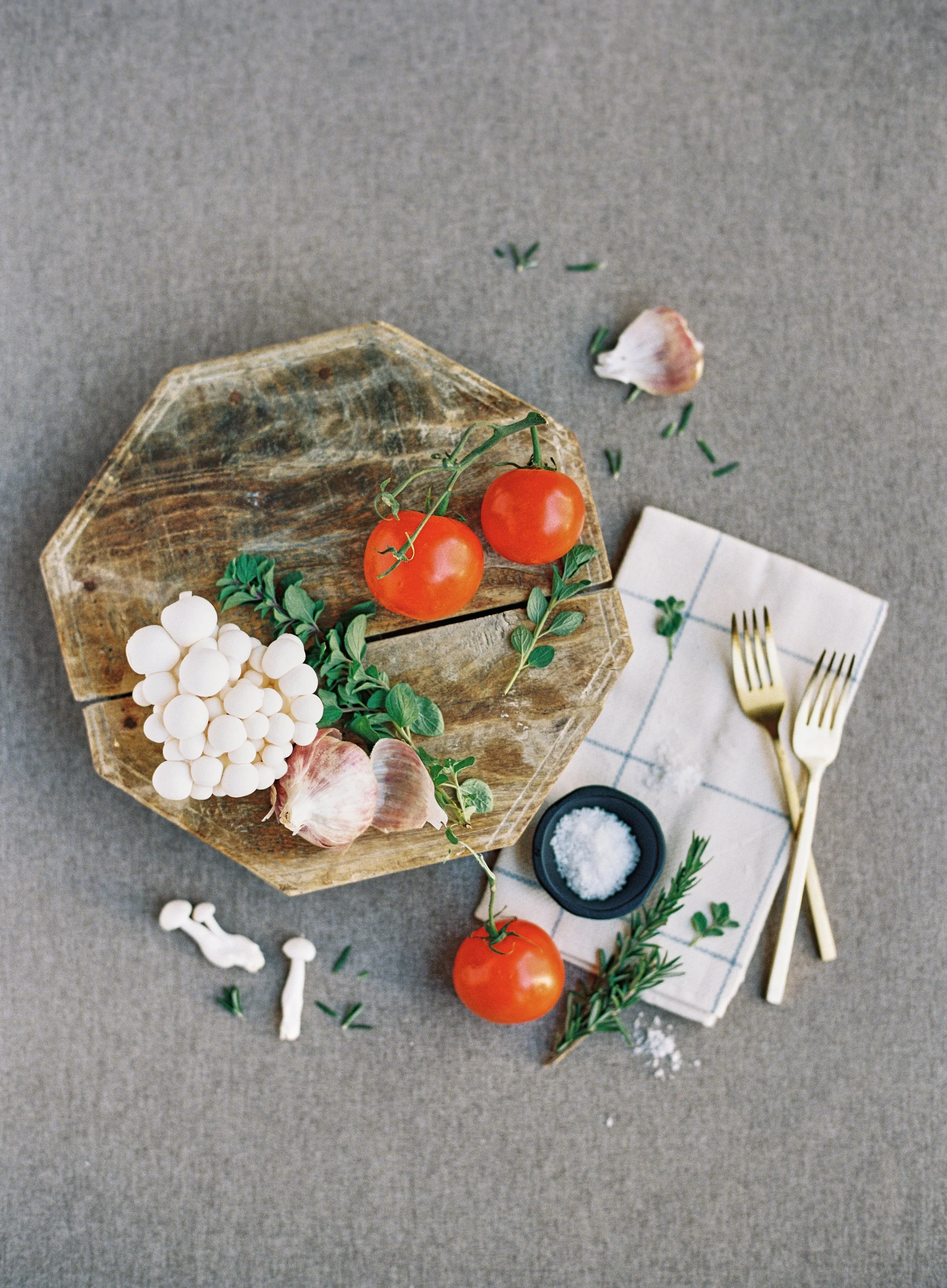 food flat lay with vine tomatoes, garlic, mushrooms, and fresh herbs on a rustic wooden board styled on a cool grey hand-painted surface