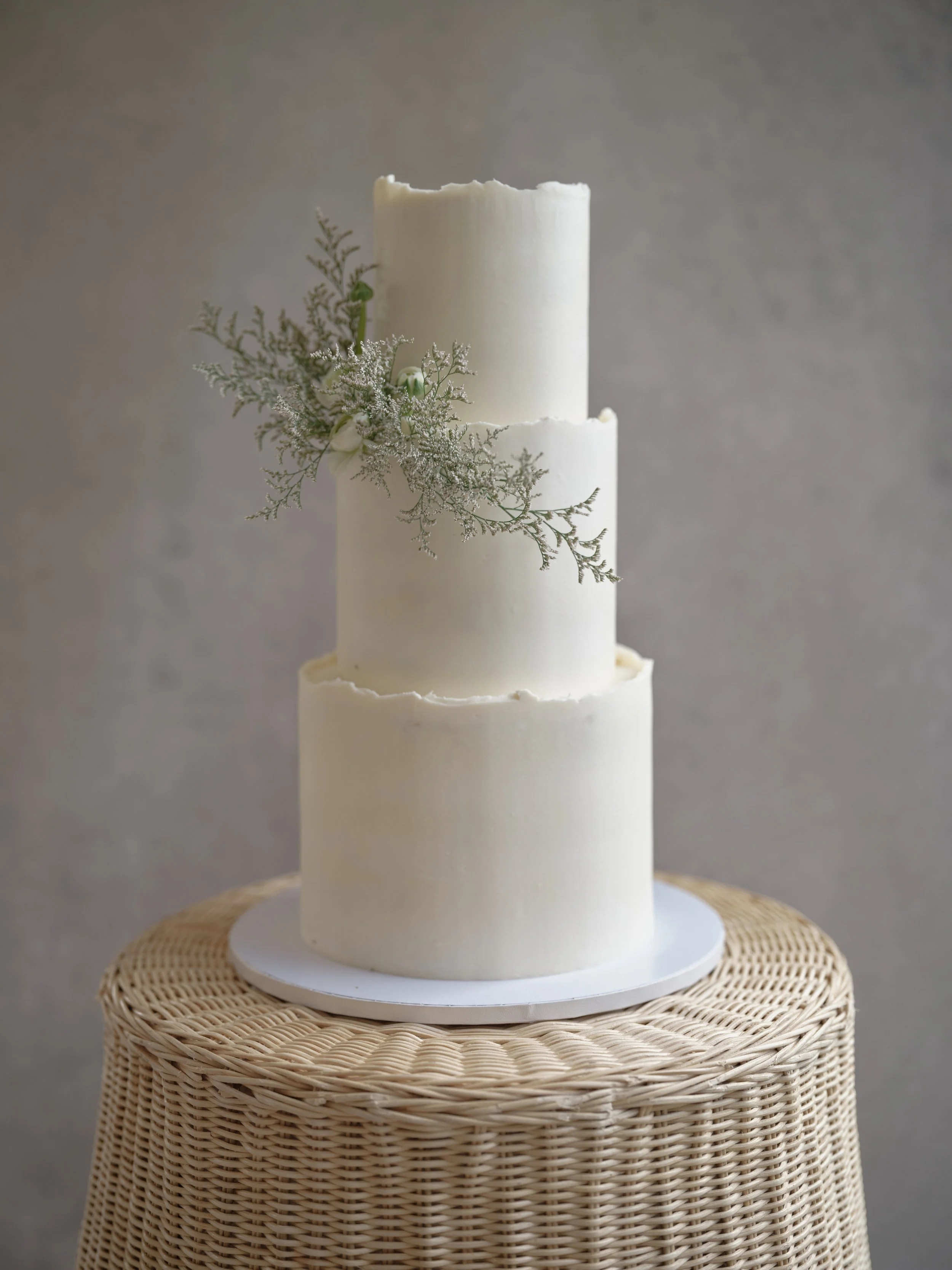 three-tier white wedding cake with deckled edges and dried floral accent on a woven wicker pedestal against a soft gray hand-painted canvas backdrop
