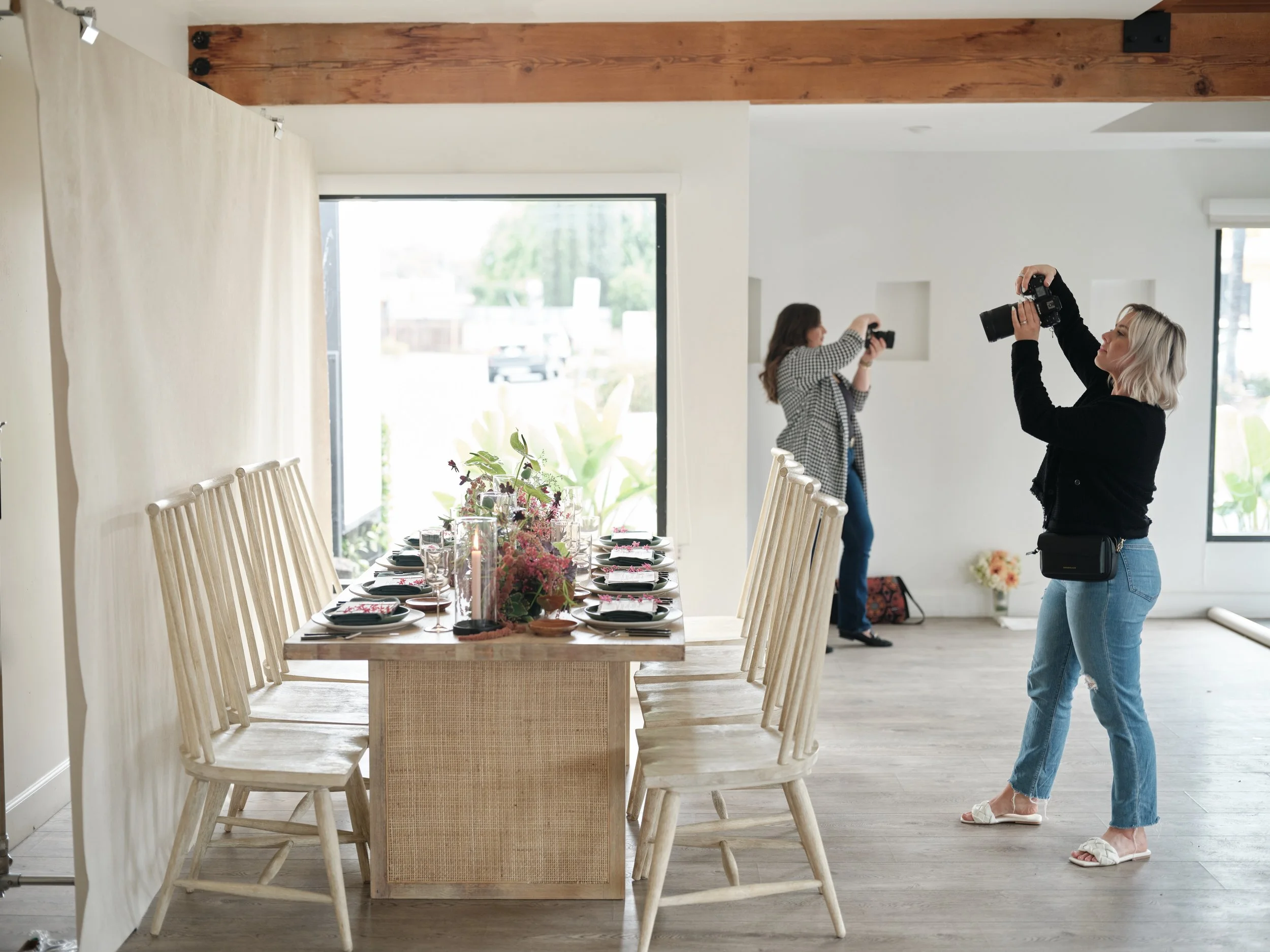 Two photographers capturing styled dining table with light wood spindle-back chairs and colorful floral centerpiece in bright modern space with exposed wooden beam and floor-to-ceiling windows