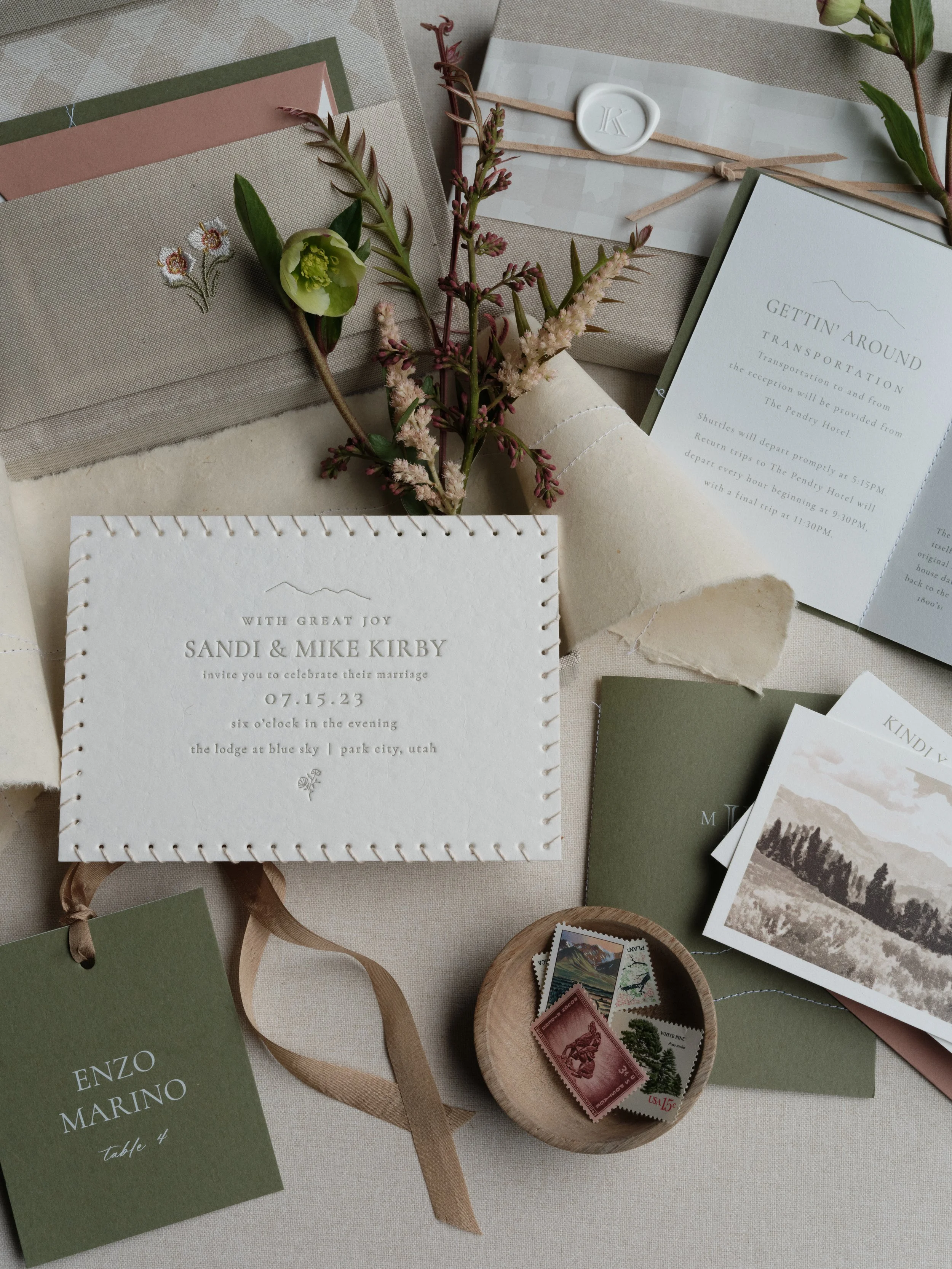 Letterpress wedding invitation suite flat lay scattered composition with sage green stationery, wax seal, vintage stamps, and botanical accents on textured photography surface