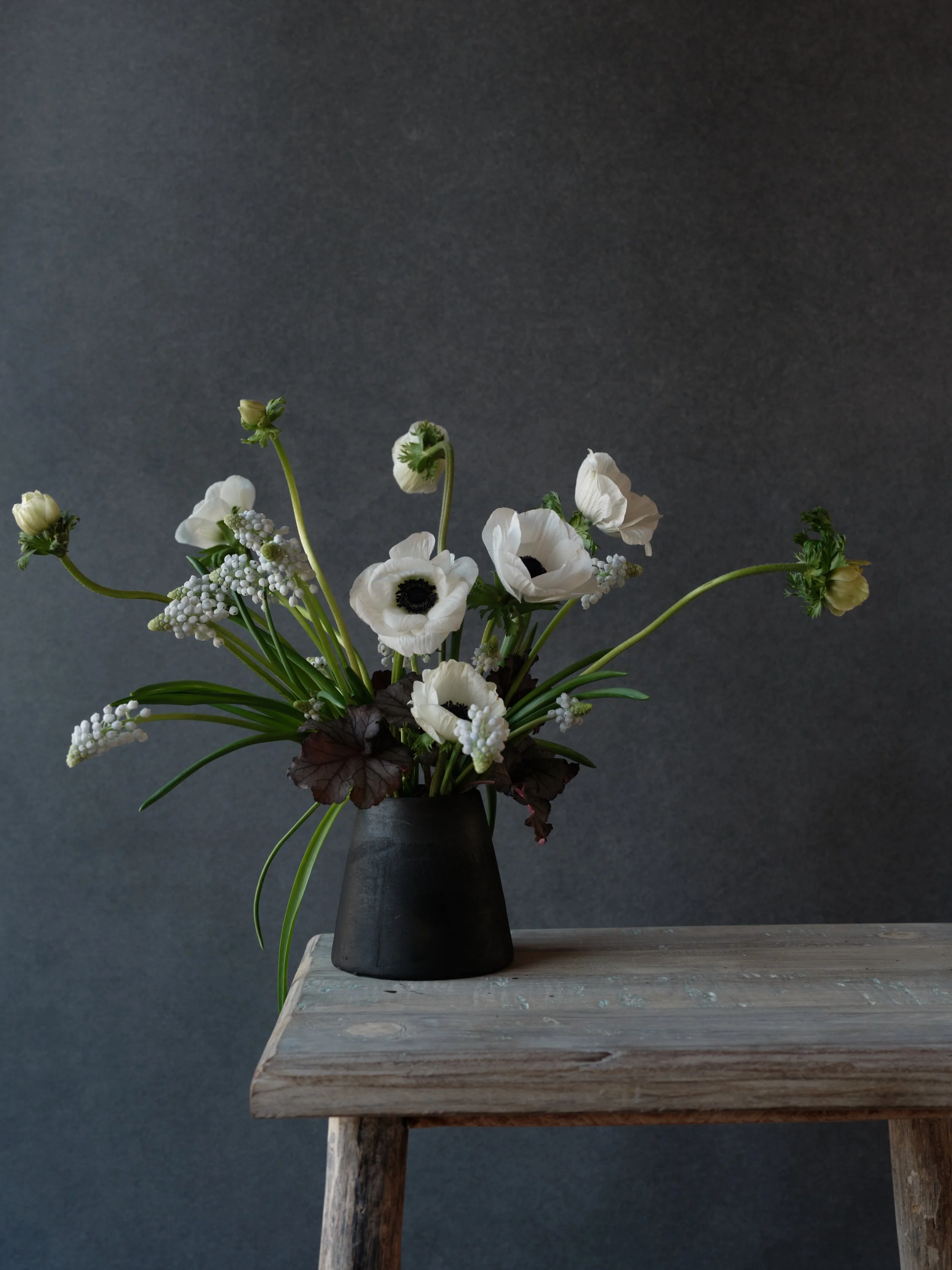 white anemone and grape hyacinth arrangement in a dark ceramic vessel on a weathered wooden bench against a Graphite hand-painted canvas backdrop
