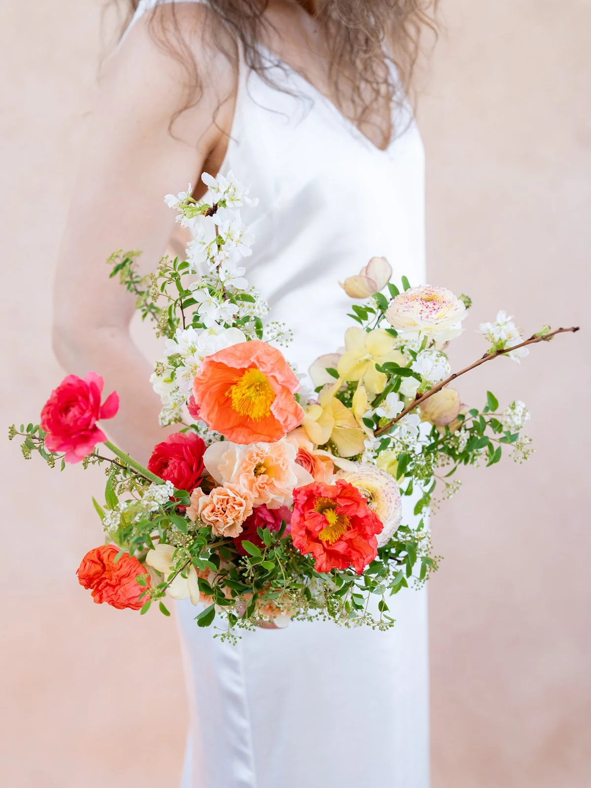 bride in white silk slip dress holding a spring bouquet of coral poppies, pink ranunculus, and white cherry blossoms in front of a Rose-Quartz hand-painted canvas backdrop