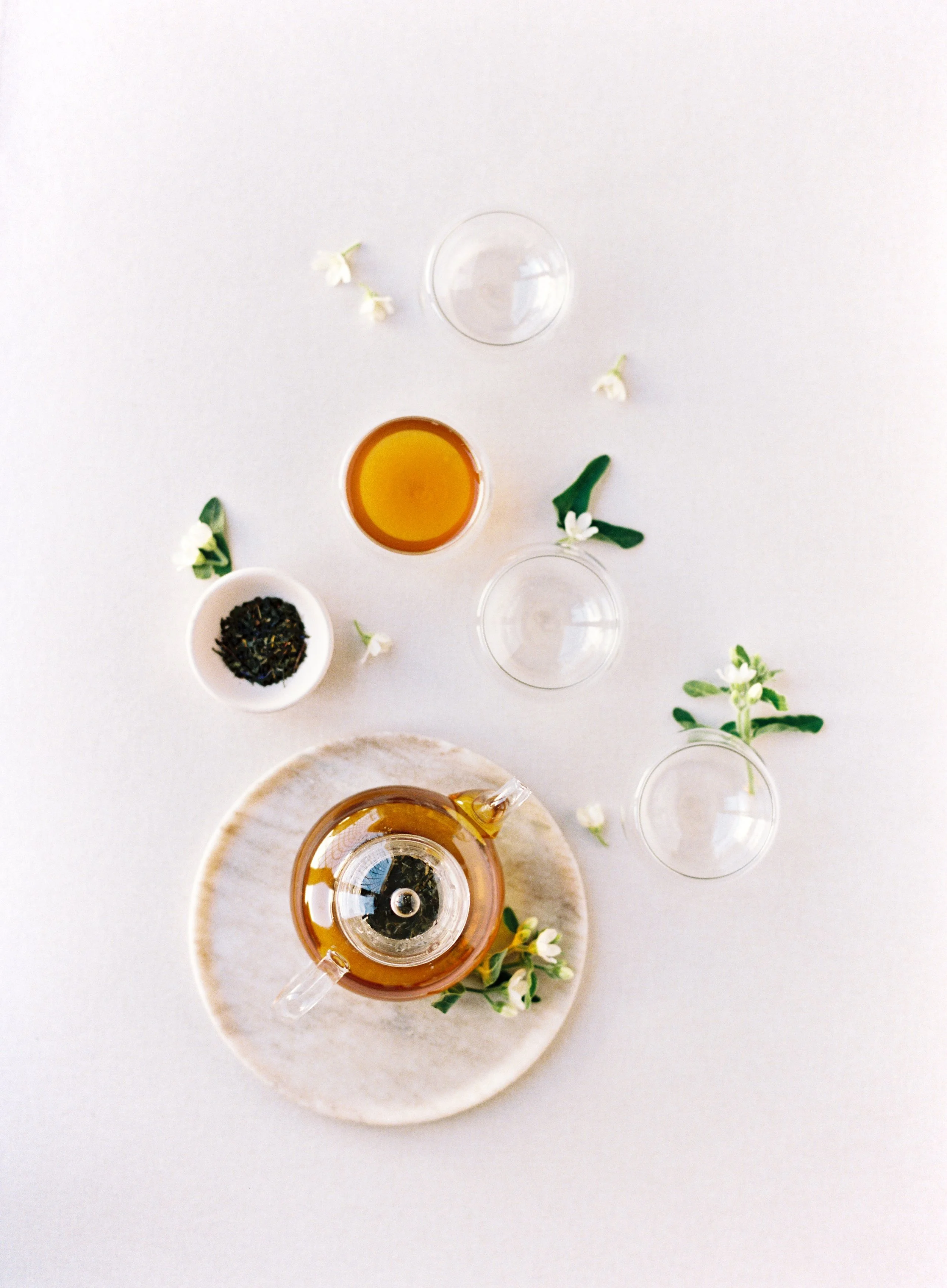 Minimalist tea flat lay photography with glass teapot, loose leaf tea, teacups, and fresh white blossoms on blush pink styling mat