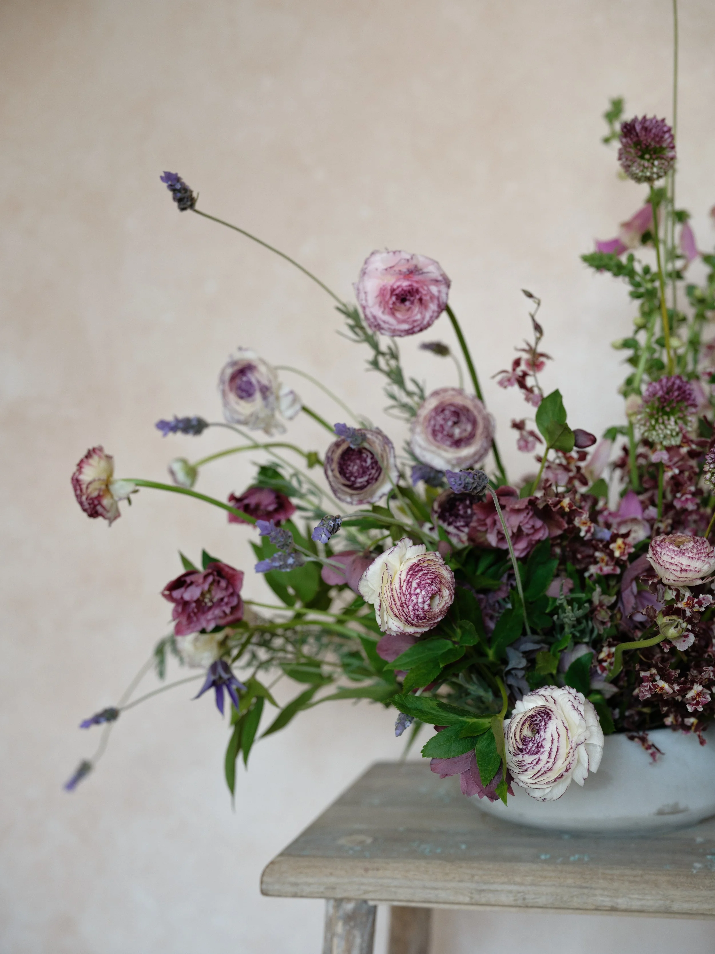 Asymmetrical floral arrangement with ranunculus and greenery styled on a rustic wooden table against a soft neutral textured backdrop