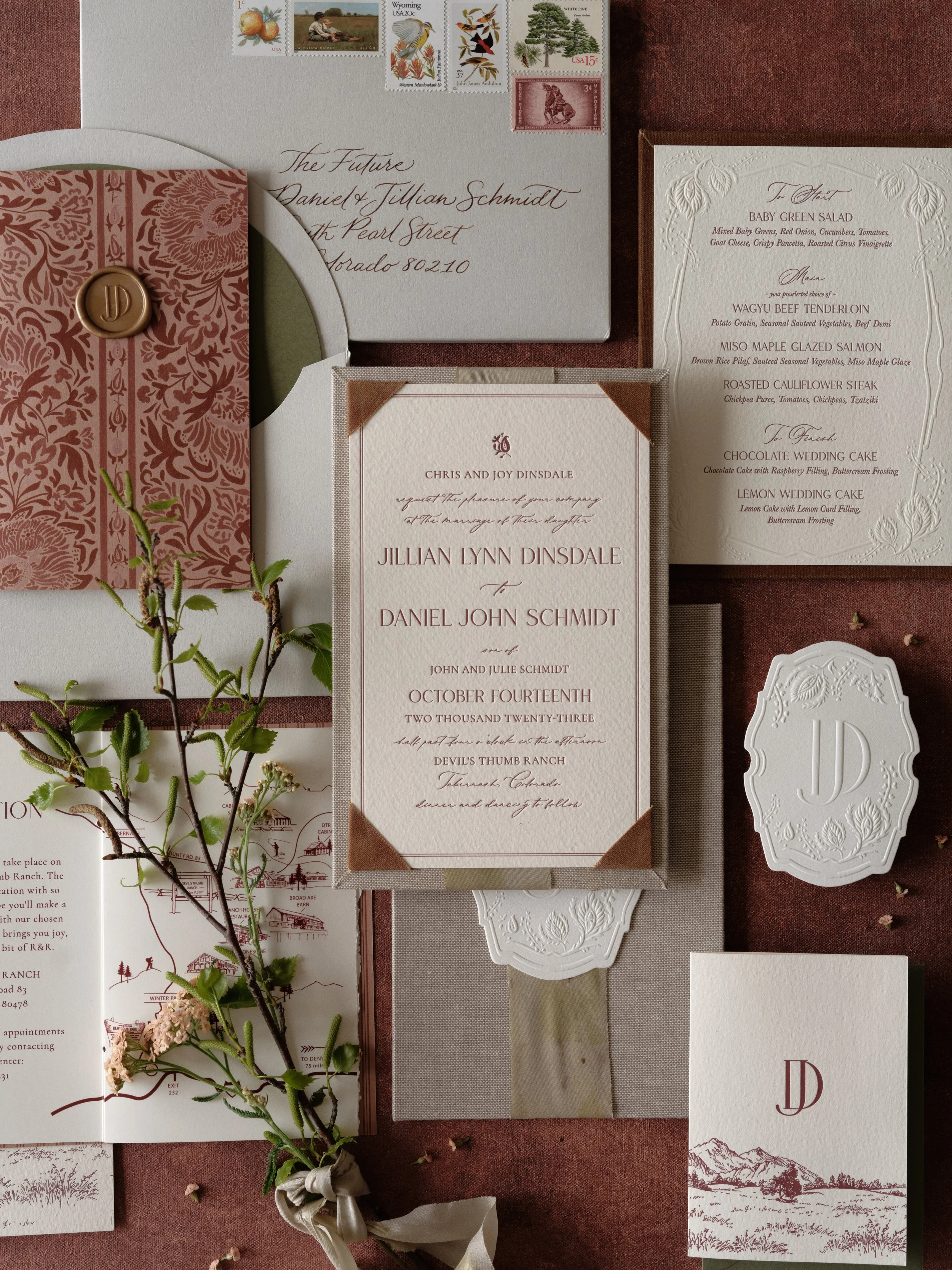 Luxury wedding invitation suite flat lay in burgundy and cream featuring embossed stationery, vintage postage stamps, calligraphy addressing, letterpress menu card, custom monogram with botanical details, and silk ribbon on textured linen backdrop