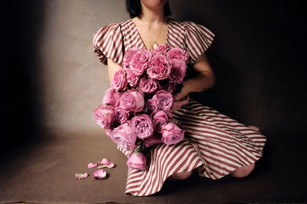 Woman in a striped dress holding a large bouquet of pink garden roses against a hand-painted canvas backdrop, showcasing luxury portrait photography with soft editorial styling