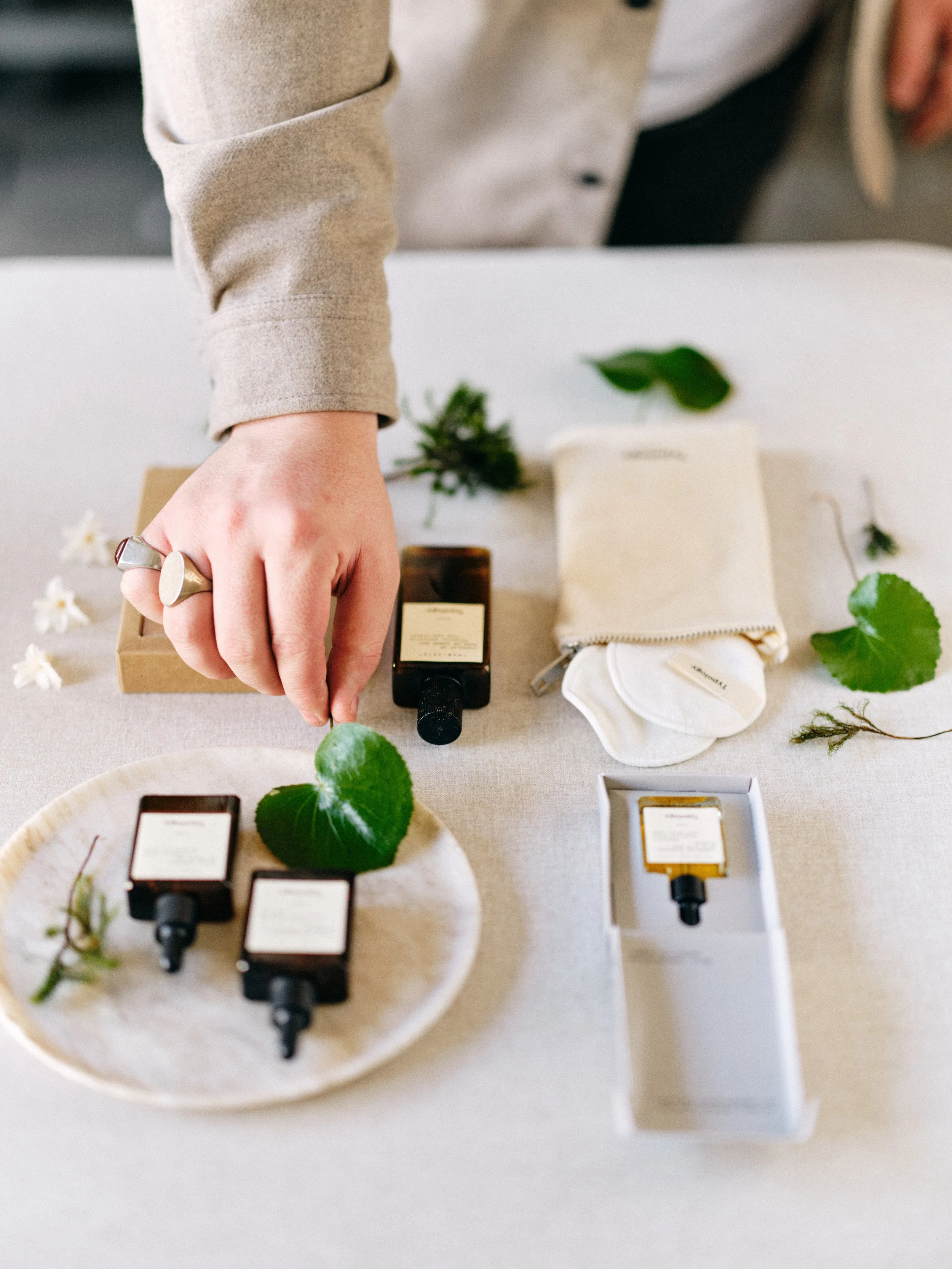 Hands styling a skincare product flat lay with amber bottles, botanical accents, and packaging on a neutral hand-painted surface