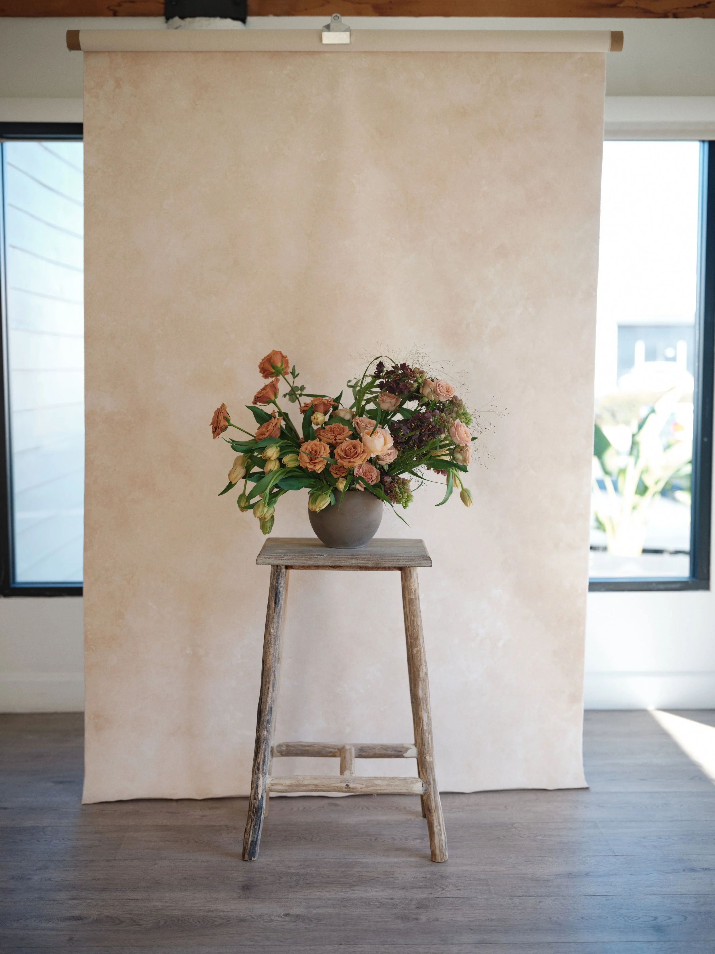 Floral arrangement in a ceramic vase placed on a wooden stool against a full-length hand-painted neutral canvas backdrop in a natural light studio