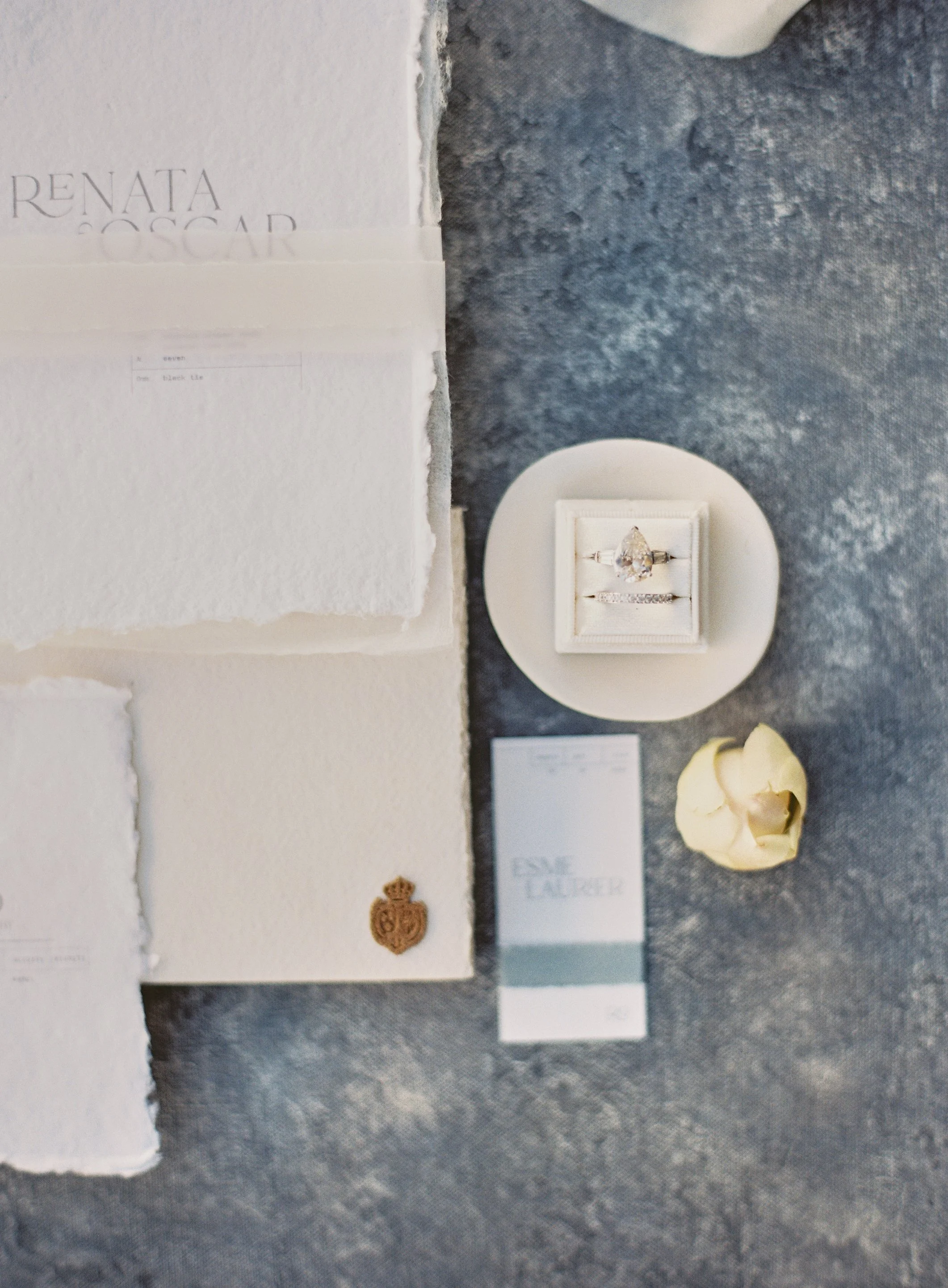 Minimal frame wedding flat lay with pear diamond ring, deckled edge stationery, gold wax seal, and cream flower on textured blue photography surface