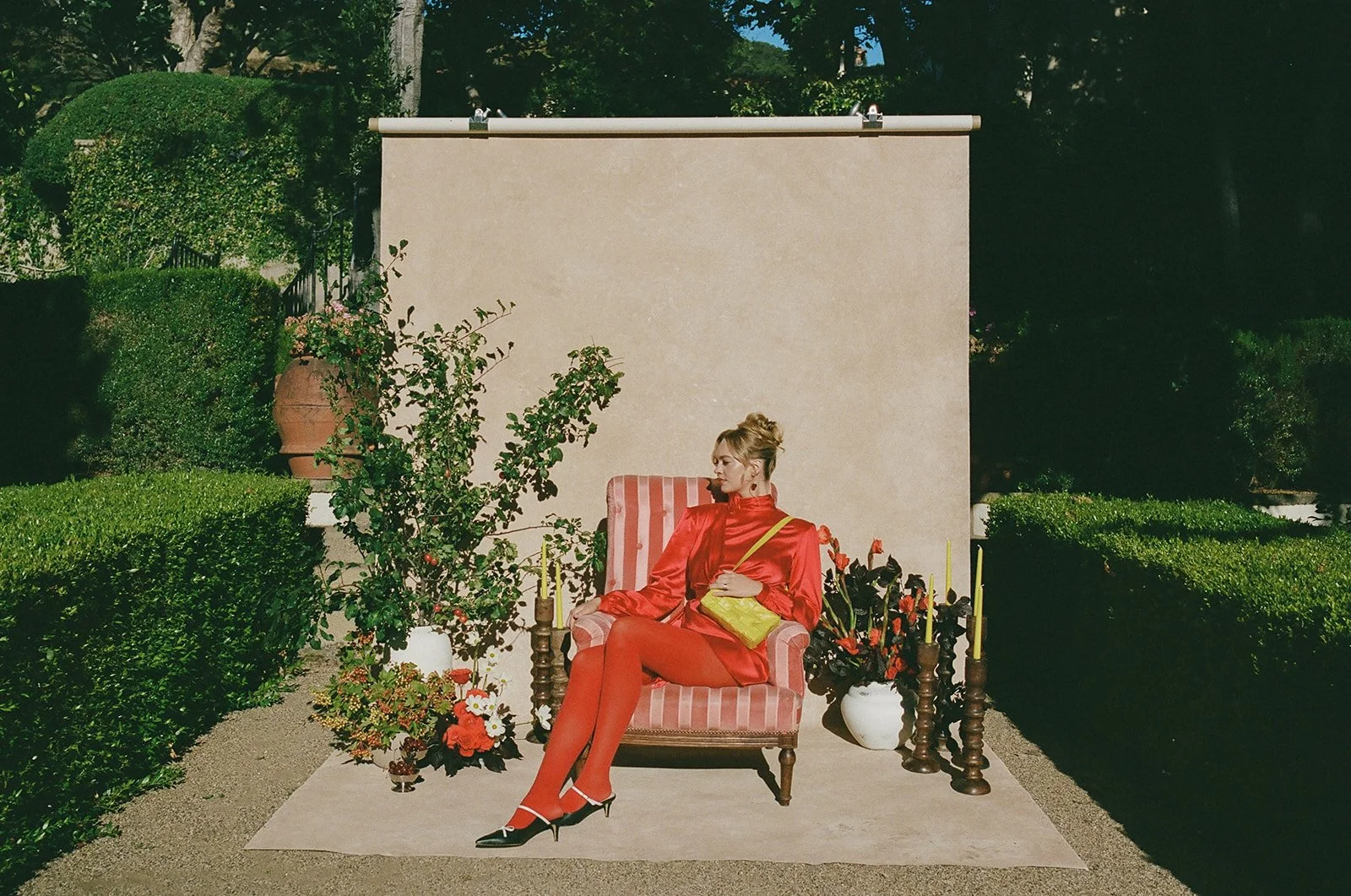 Outdoor editorial portrait of a model in a red satin ensemble seated in a striped armchair against a Chasing Stone Mica canvas backdrop in a garden setting