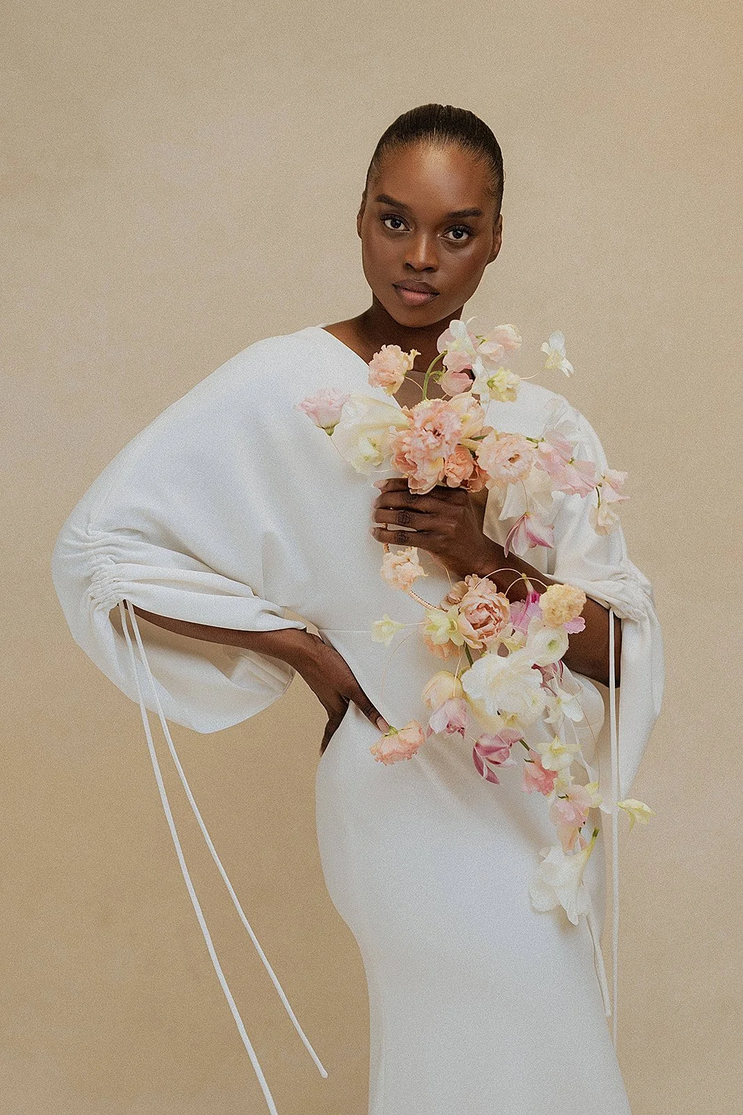 bridal fashion editorial portrait of a model in a white draped gown with ruched drawstring sleeves holding a cascading peach and pink floral arrangement against a neutral beige backdrop