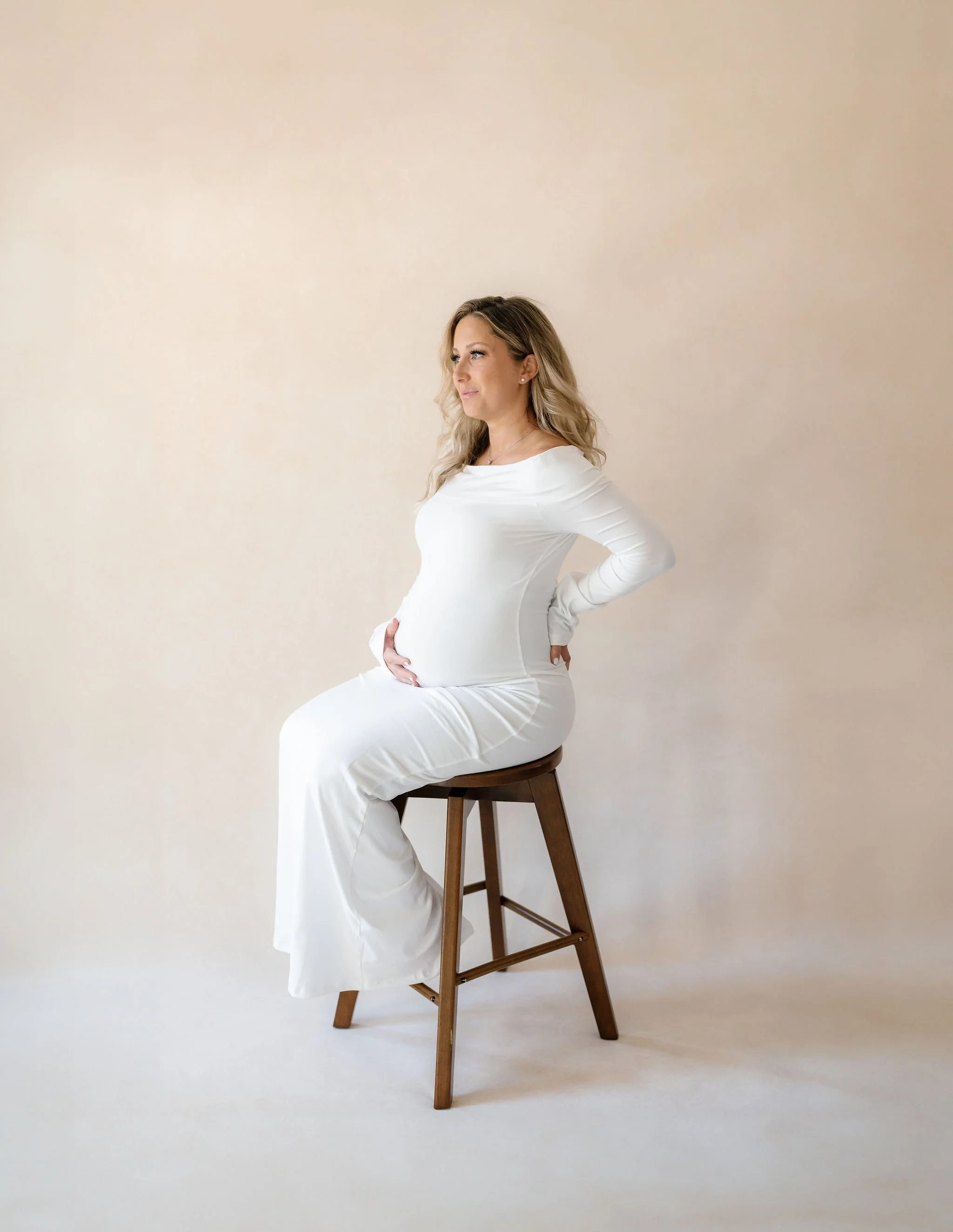 pregnant woman in a white maternity gown sitting on a stool against a soft neutral studio backdrop.