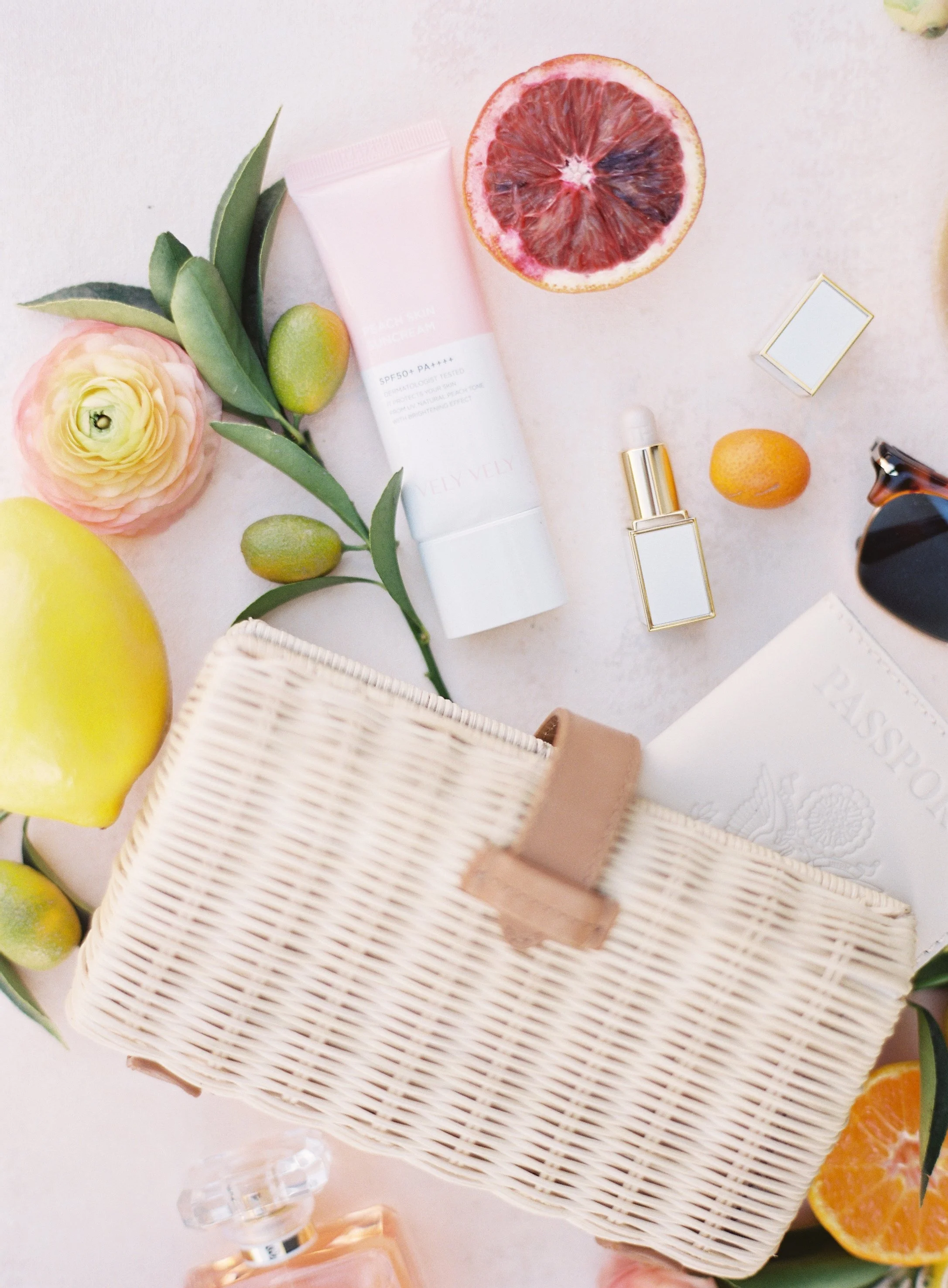 Bright and airy flat lay of beauty products and citrus fruits on warm blush pink hand-painted styling surface with soft directional lighting and organic floral elements