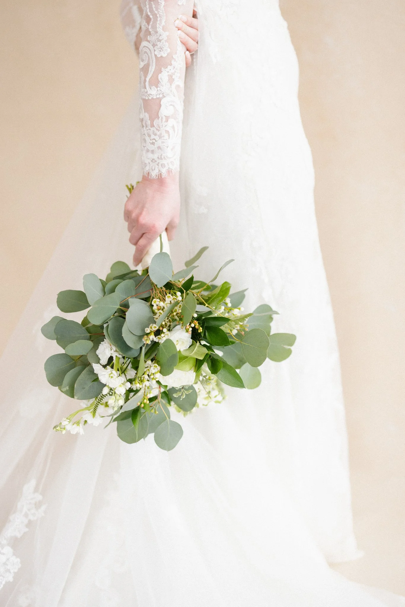 bride in long-sleeve lace gown holding a eucalyptus and white rose bouquet against a warm neutral canvas backdrop