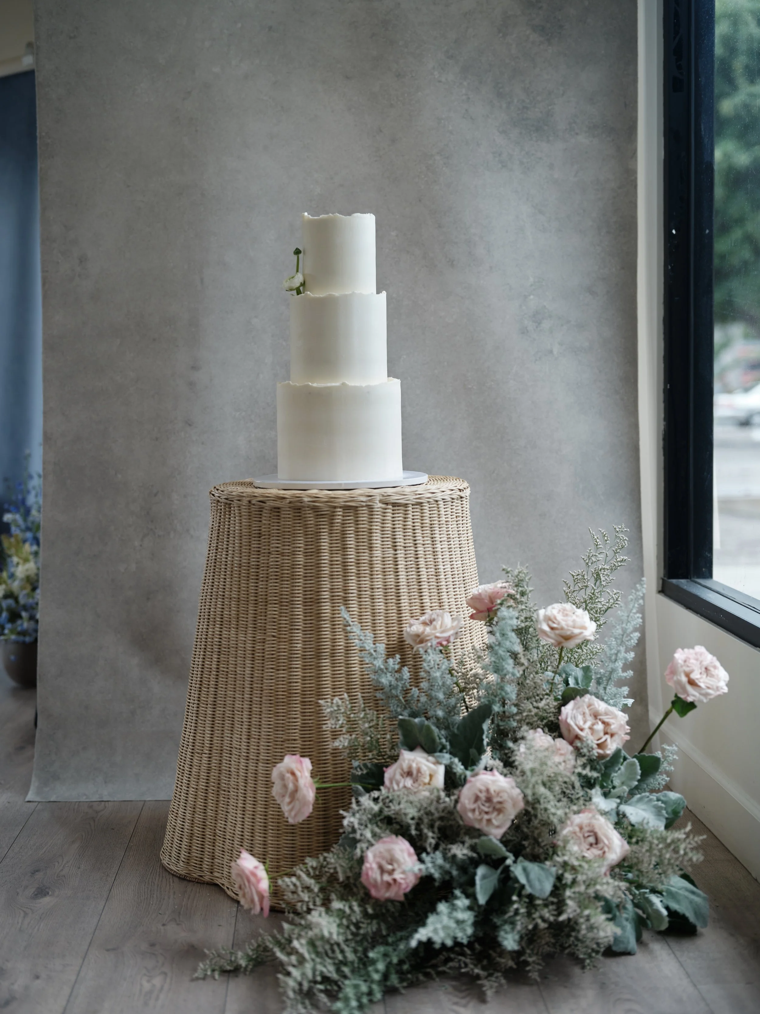 three-tier white wedding cake on woven stand with pink roses and eucalyptus styled on Chasing Stone travel-inspired hand-painted canvas backdrop