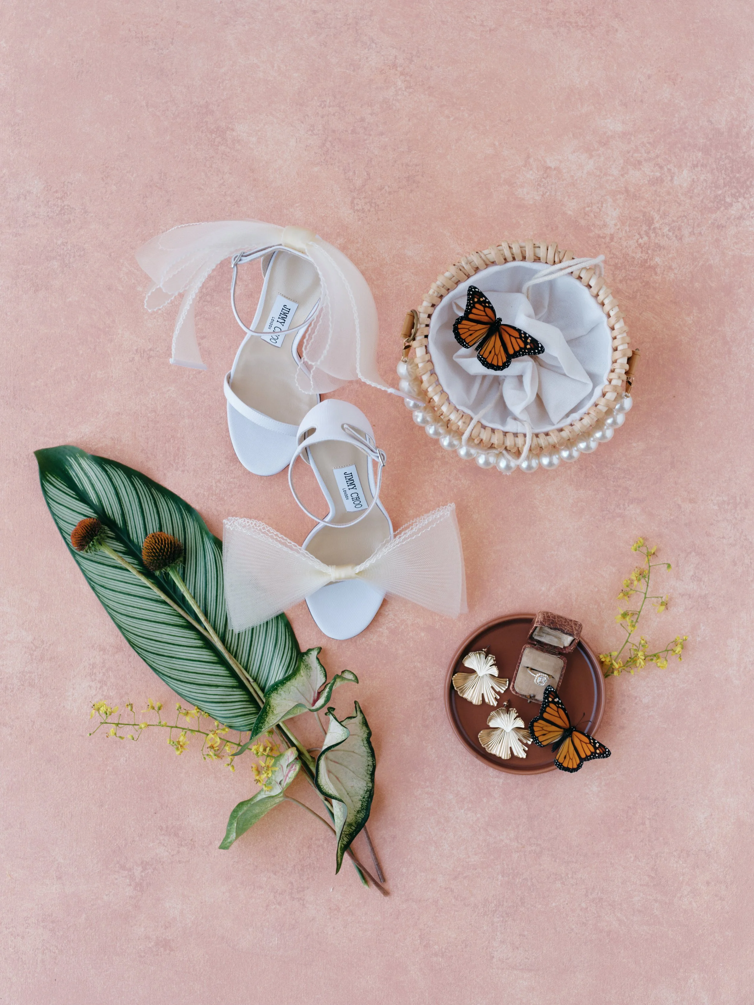 Bridal flat lay scattered composition with Jimmy Choo heels, monarch butterfly details, and ring box on pink hand-painted photography surface