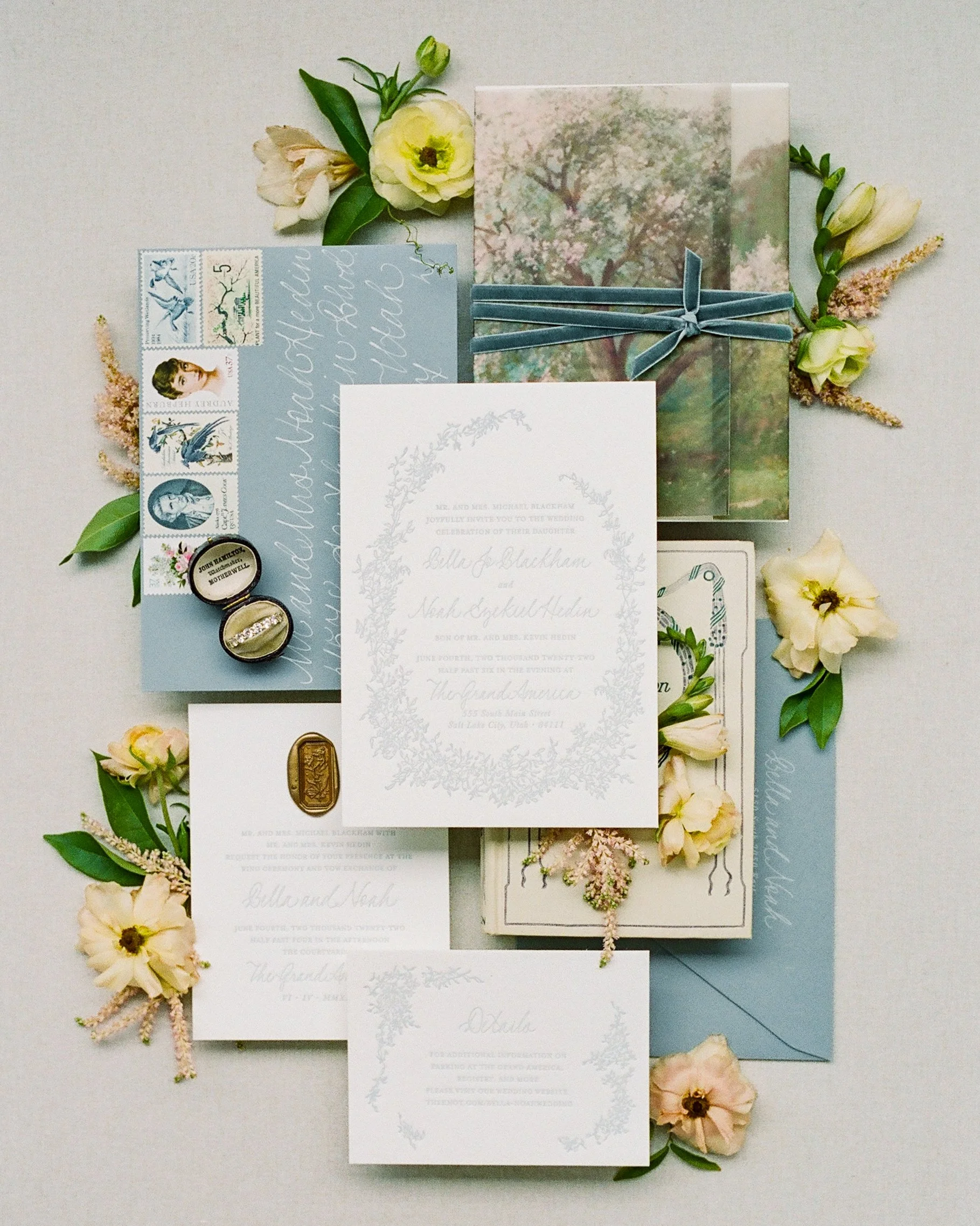 Wedding flat lay photography showing layered composition with invitation suite, vintage stamps, velvet ribbon, and florals on alder blue-gray styling surface