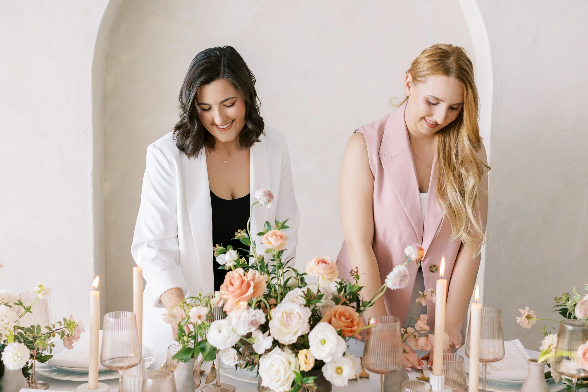 Two women styling an elegant floral wedding tablescape with candles and soft natural light in an editorial event setting