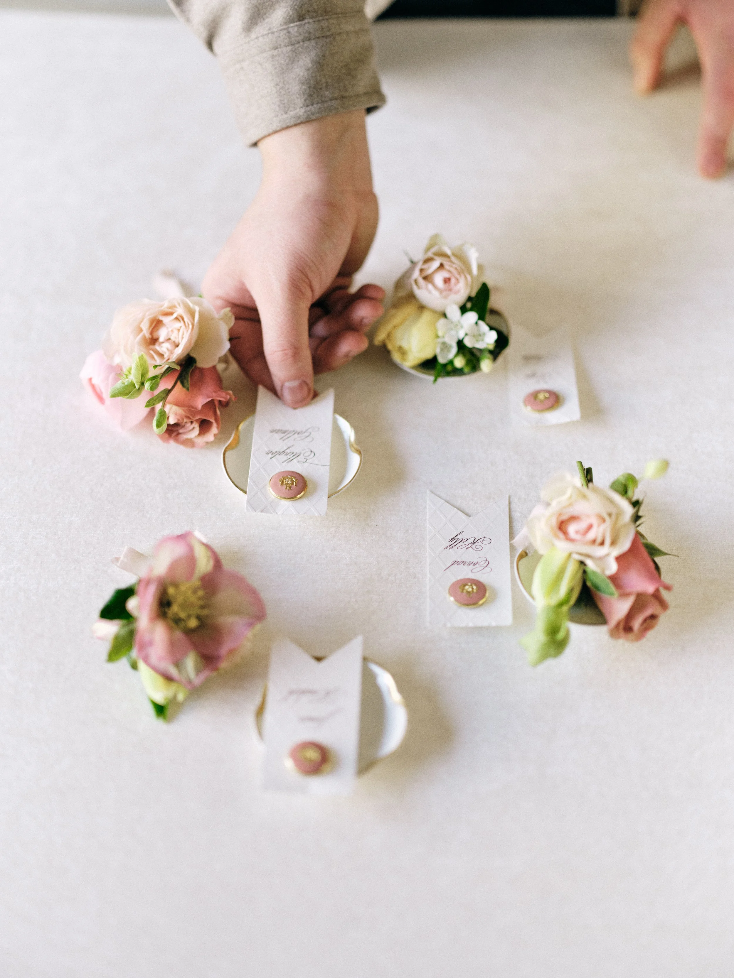 "Hand arranging delicate boutonnières with blush and cream roses, hellebores, and green accents on neutral surface alongside calligraphy place cards with wax seals