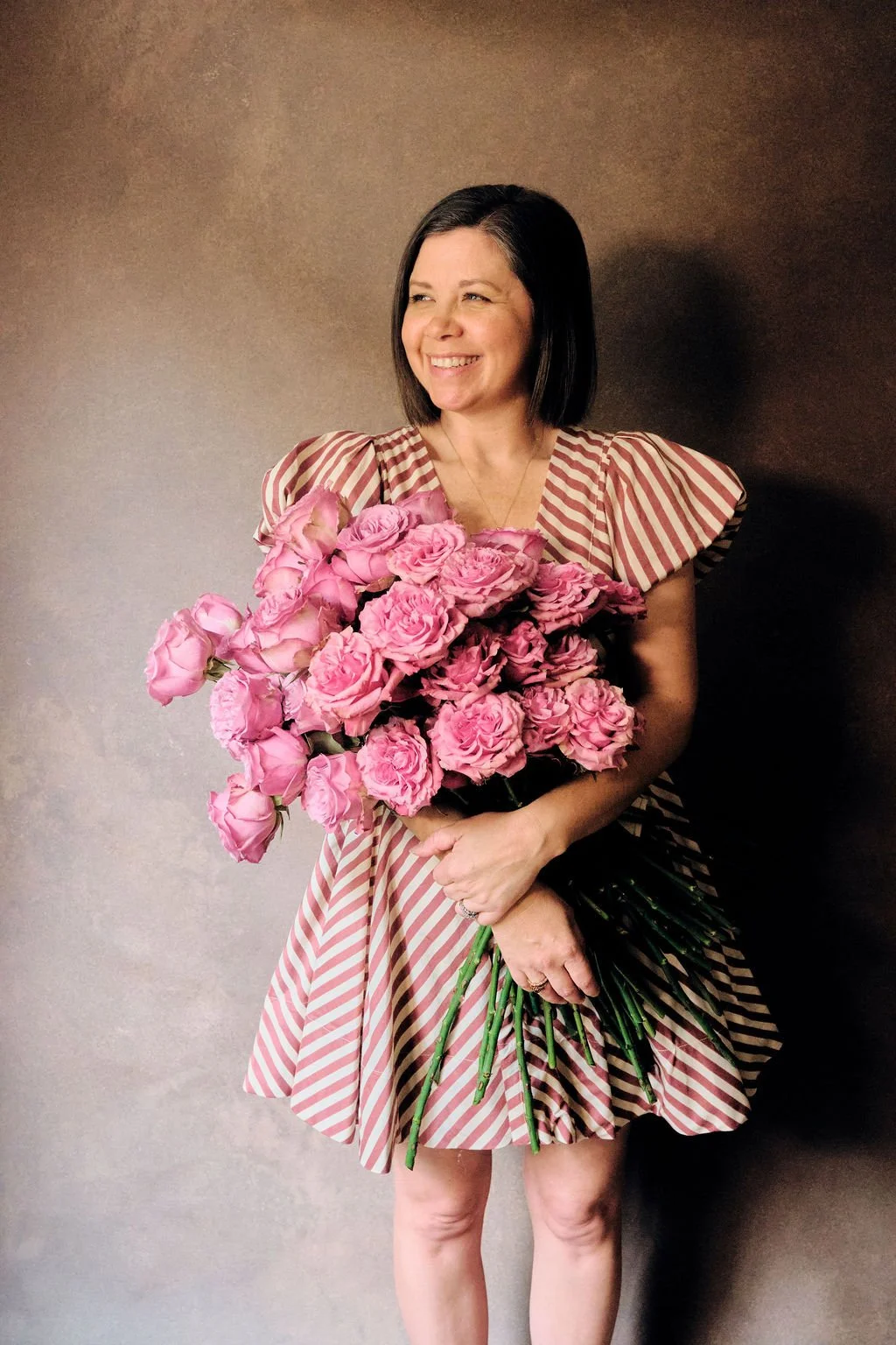 Smiling woman in a pink and white striped dress holding a large bouquet of pink roses against a warm-tone hand-painted canvas backdrop.