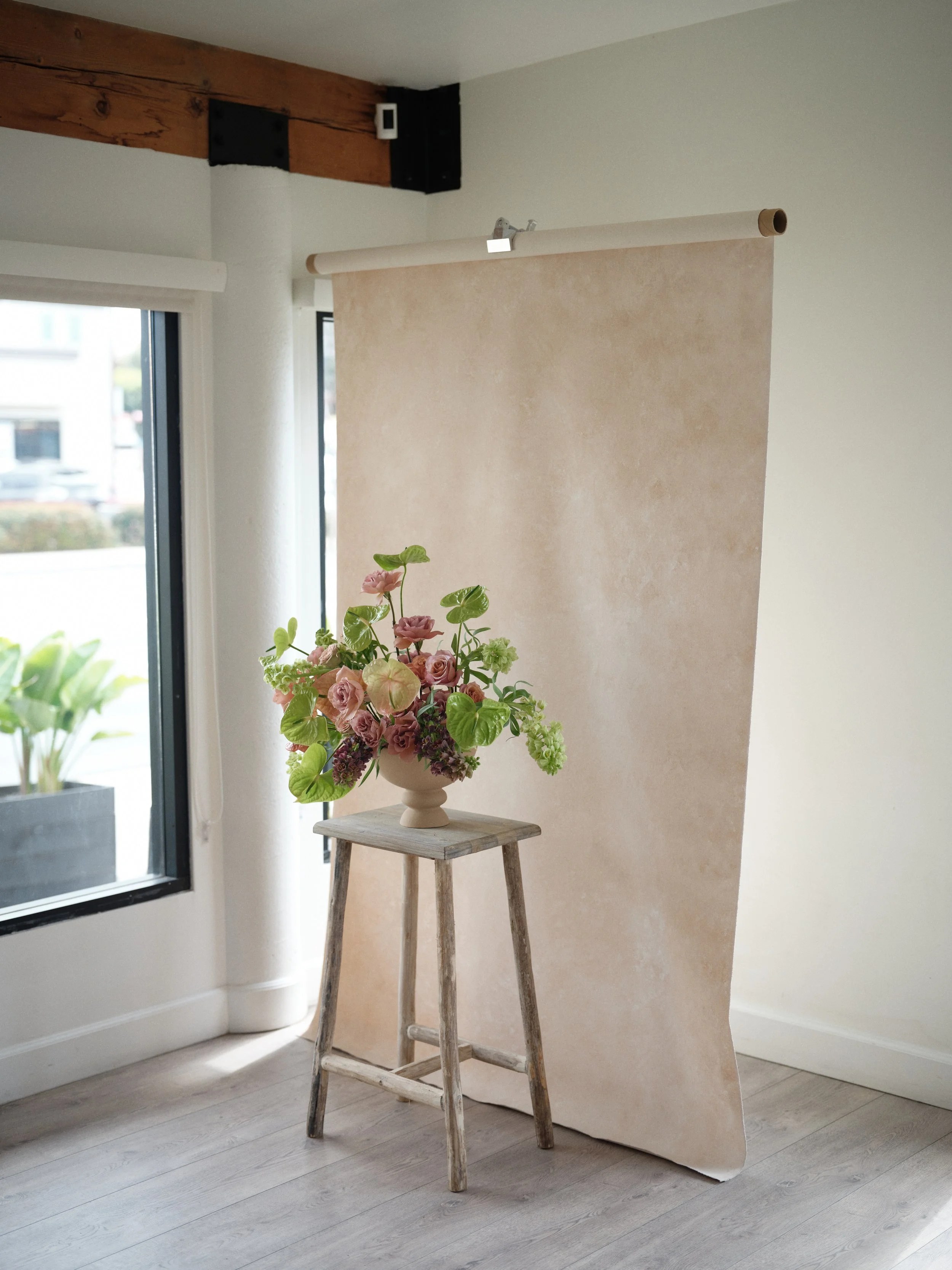 hand-painted beige canvas photography backdrop lit with soft natural window light, featuring a floral arrangement on a wooden stool in a bright studio setting