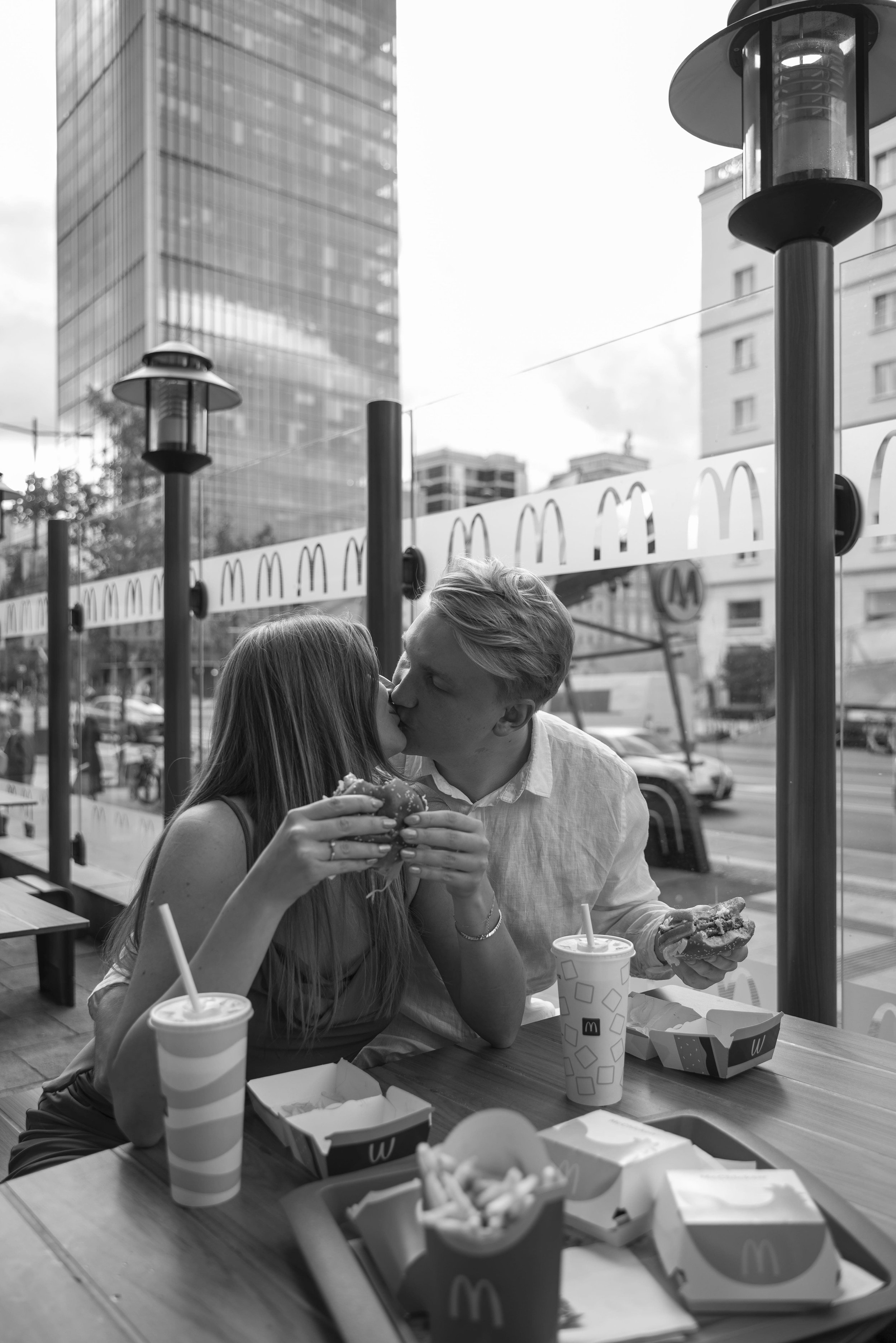 A couple sitting outside a McDonald's restaurant, sharing a kiss while holding burgers, with McDonald's packaging and fries on the table. A cityscape with tall buildings is visible in the background.