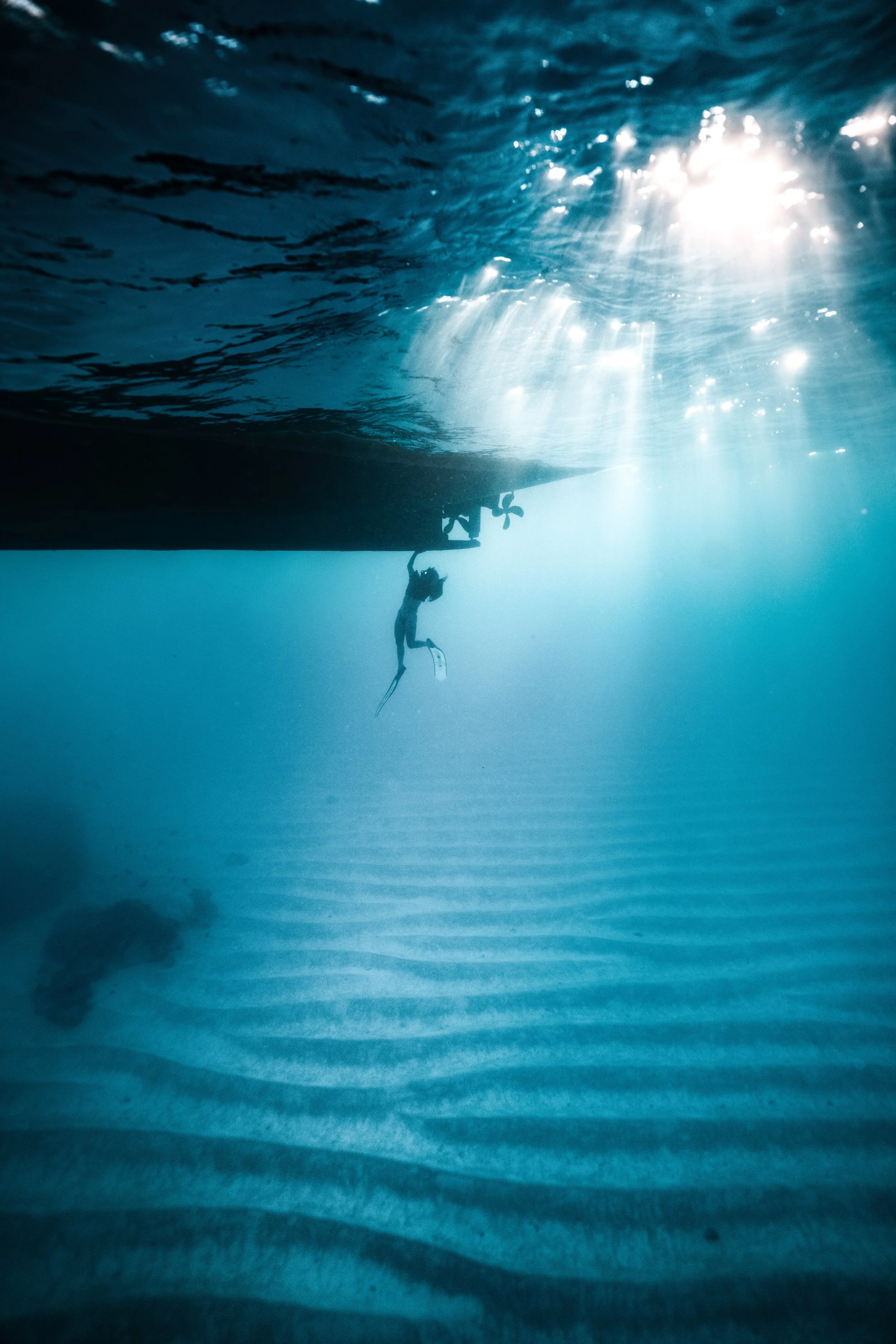 A freediver beneath a boat in clear blue water, with sunlight reaching the seabed