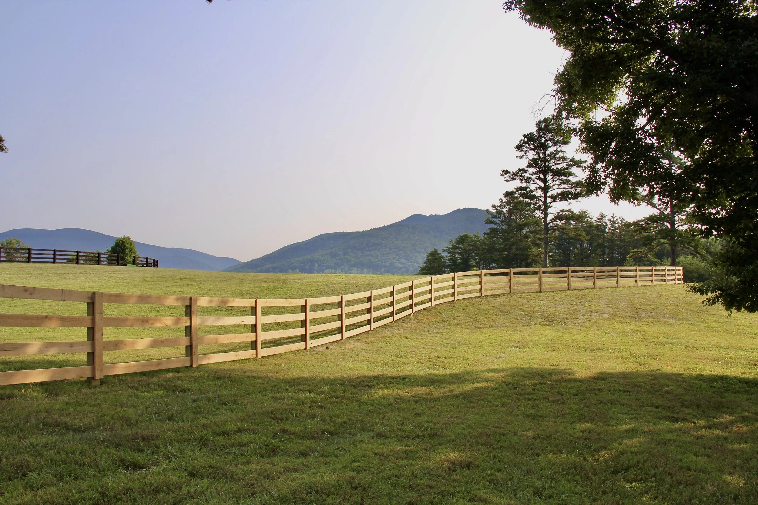 A lush green field with a wooden fence, rolling hills, and a clear sky in the background.