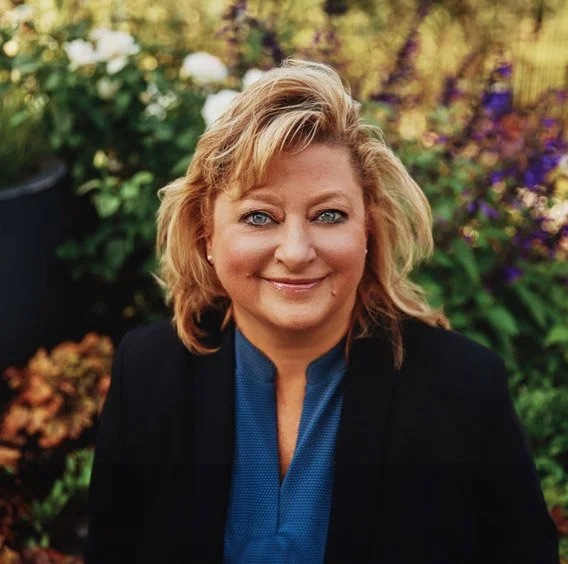 A woman with blonde hair smiling outdoors in front of colorful flowers and greenery.