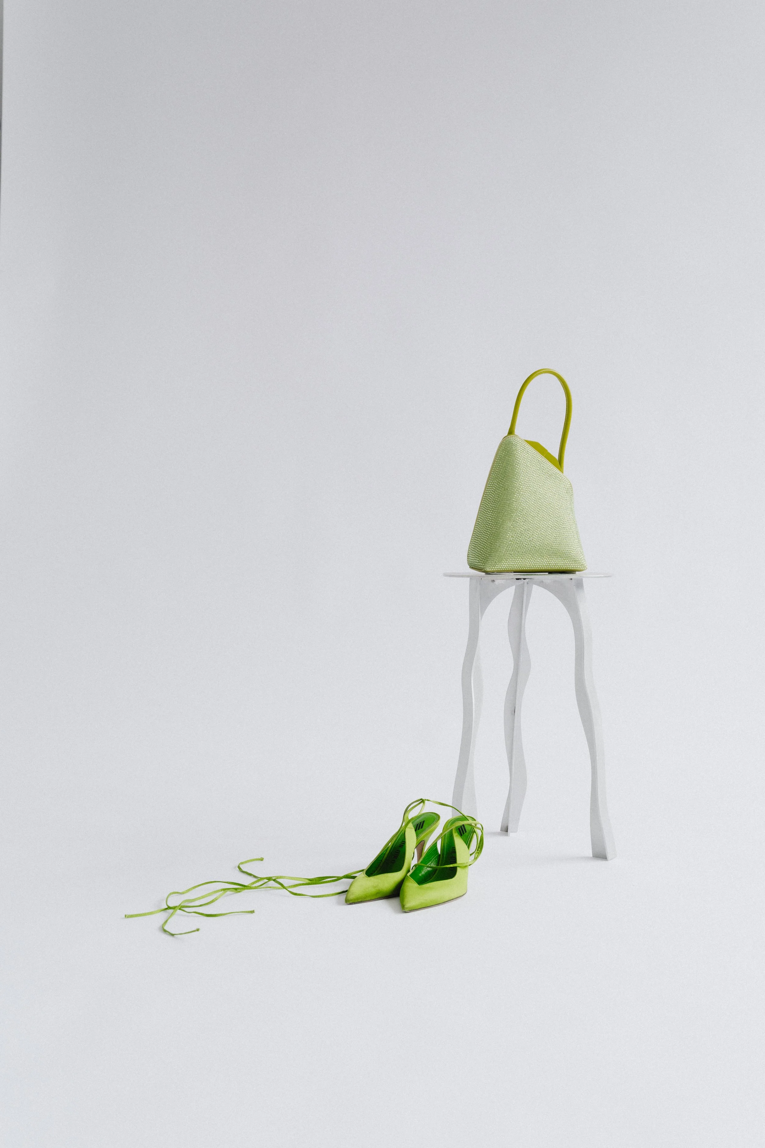 A green handbag on a white chair with white legs, and a pair of green high-heeled shoes on the floor with laces untied.