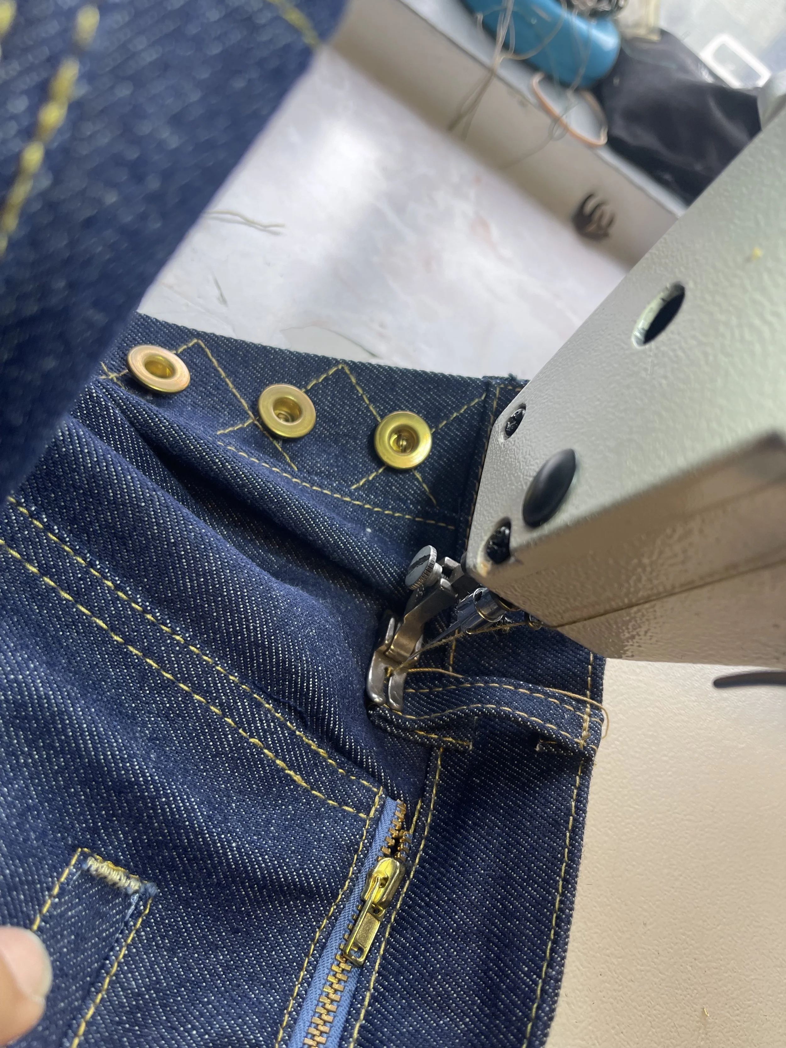Close-up of denim jeans being sewn with fabric and sewing machine, showing yellow stitching, buttons, and zipper.