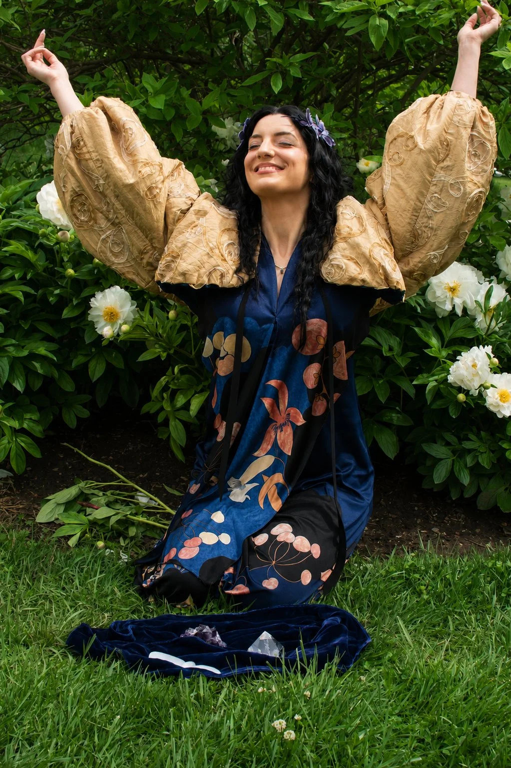 A smiling woman with long black hair, wearing a dress with a floral pattern, is kneeling on green grass in a garden with white flowers and green foliage. She has her arms raised above her head and appears to be enjoying herself.